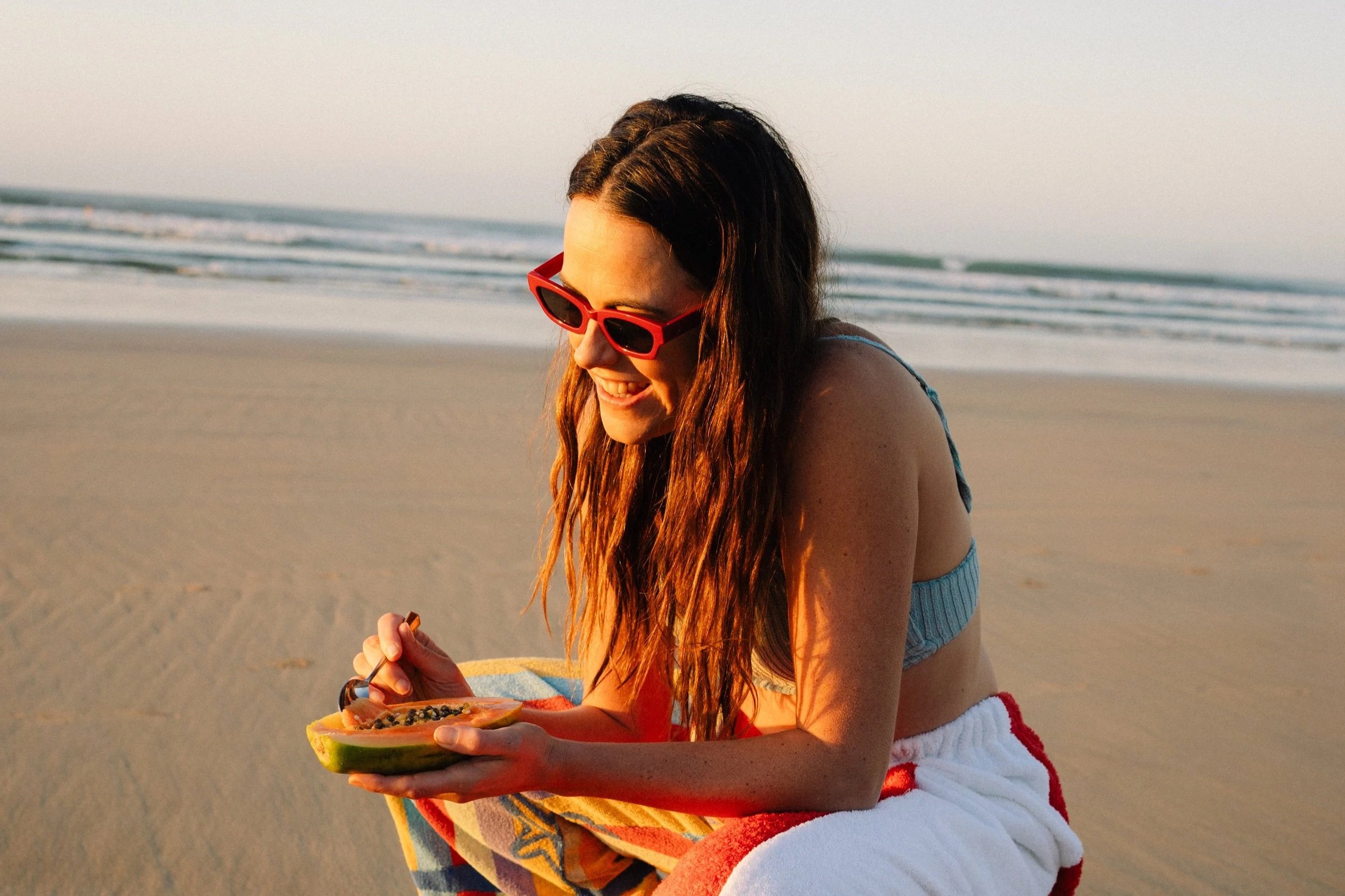 A woman with long wavy hair wearing red sunglasses and a striped swimsuit, sitting on the beach, smiling while eating fruit from a half watermelon.