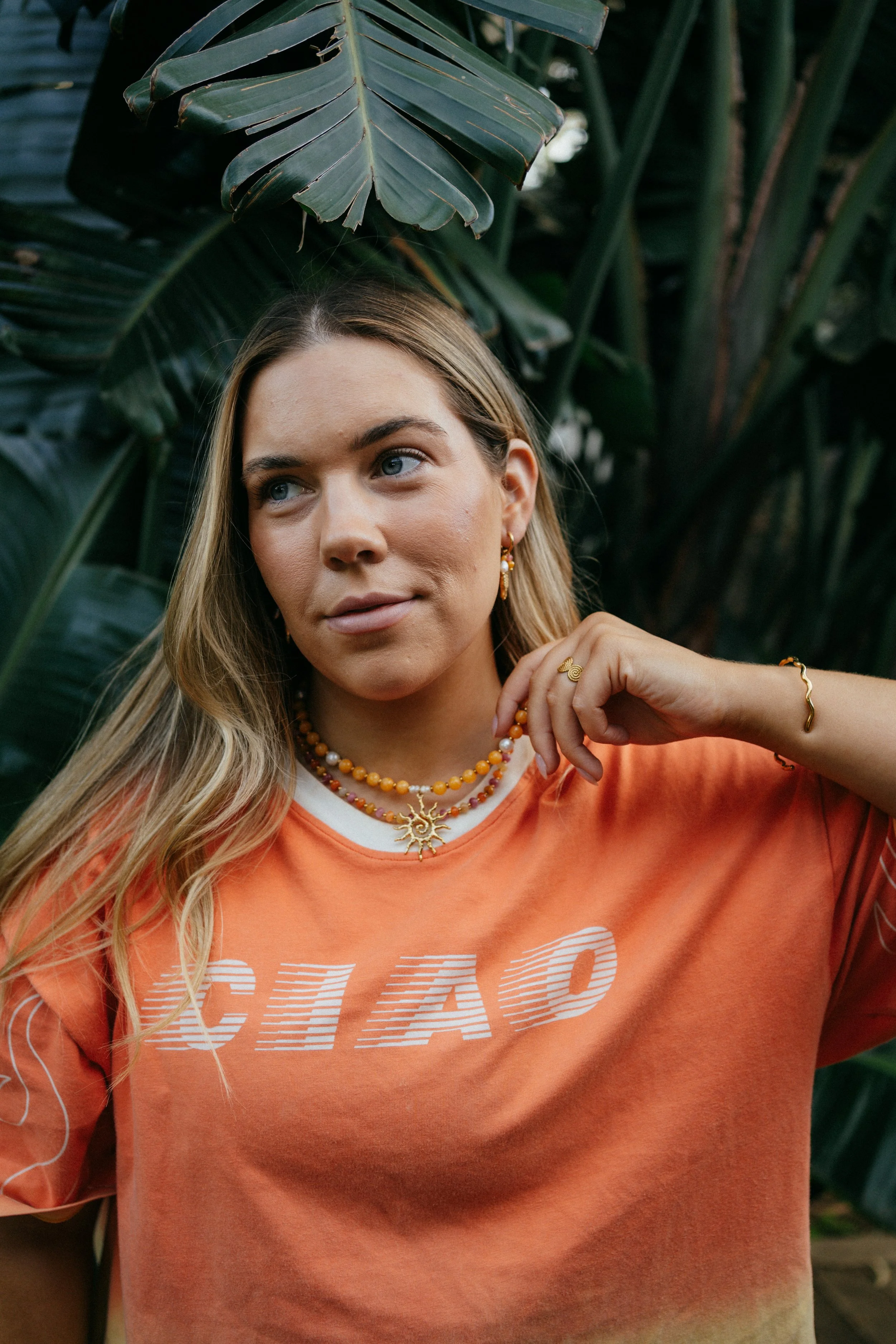 A woman with blonde hair and blue eyes wearing gold jewelry and an orange T-shirt with white text, standing in front of large green tropical leaves.