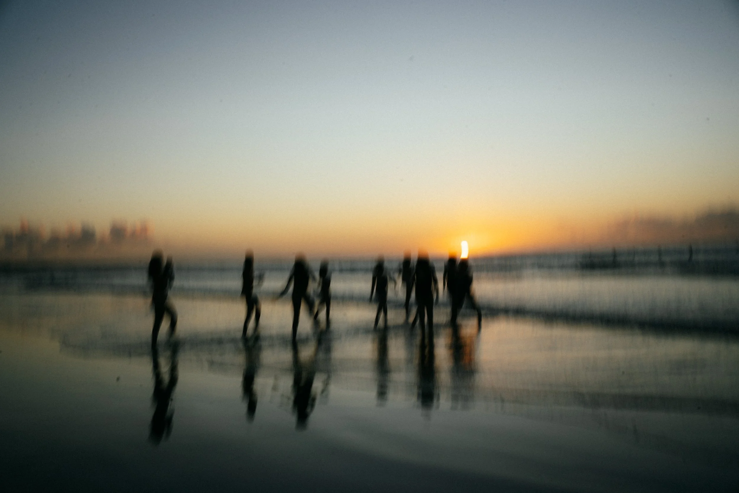 Silhouettes of people walking along the beach at sunset with a reflection in the wet sand.