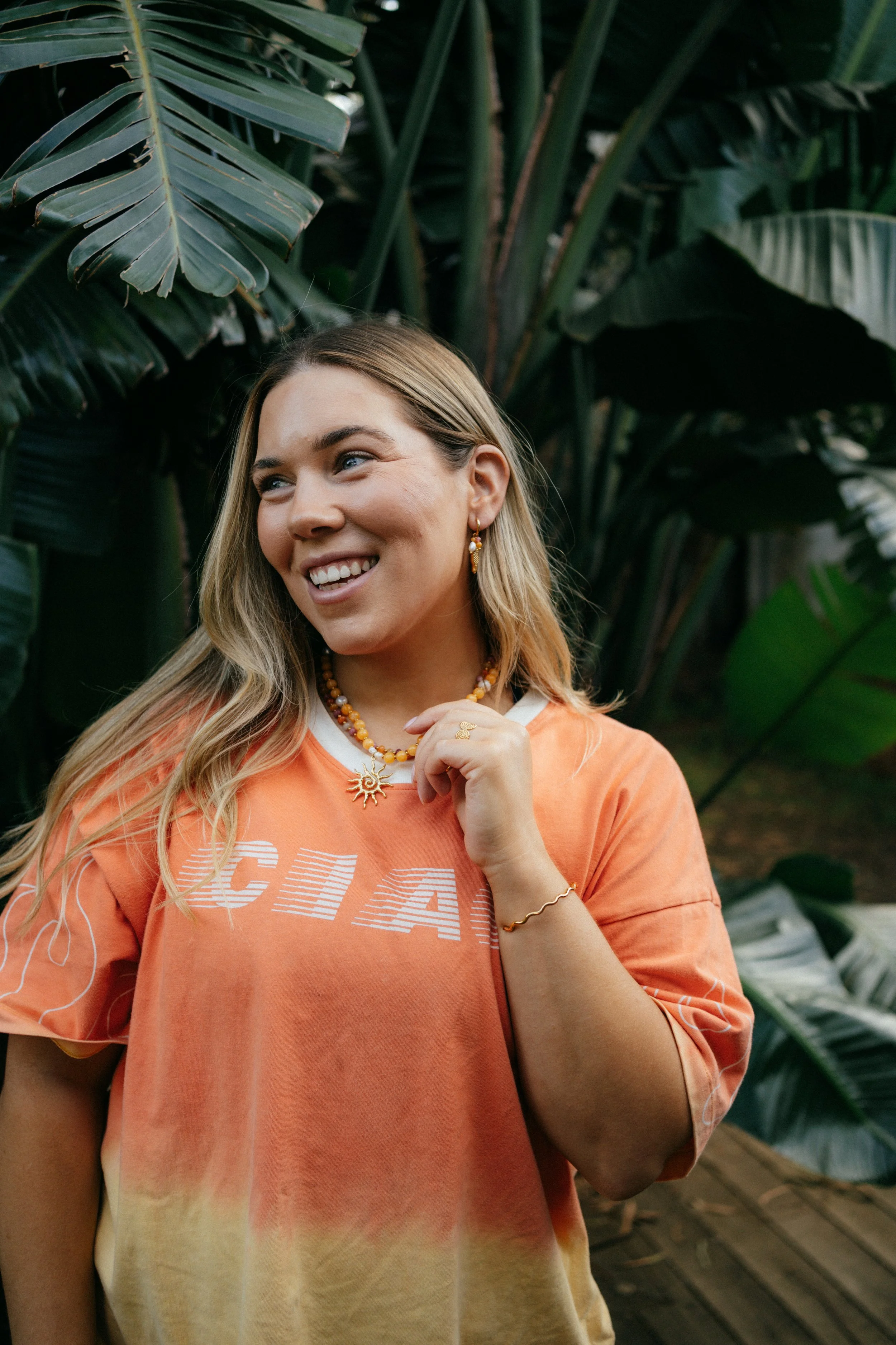 A young woman with long blonde hair wearing an orange and yellow color gradient t-shirt, gold jewelry, and smiling, standing among large green tropical leaves.