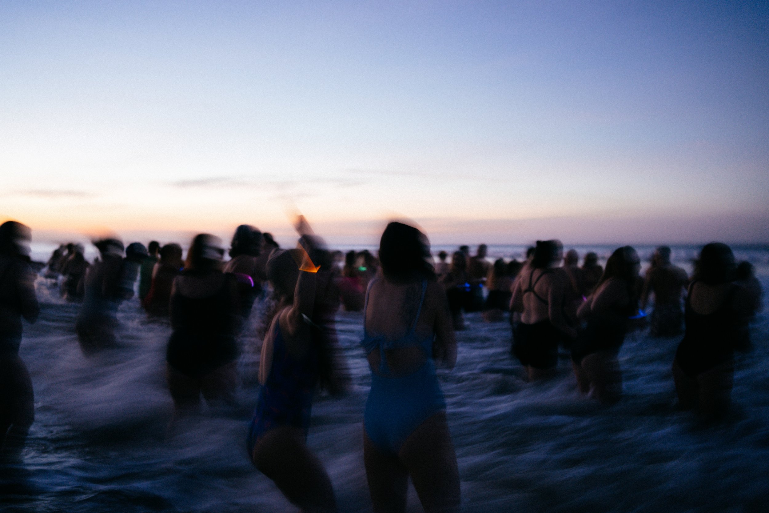 People gathered on the beach at sunset, facing the ocean with the sky transitioning from day to night.