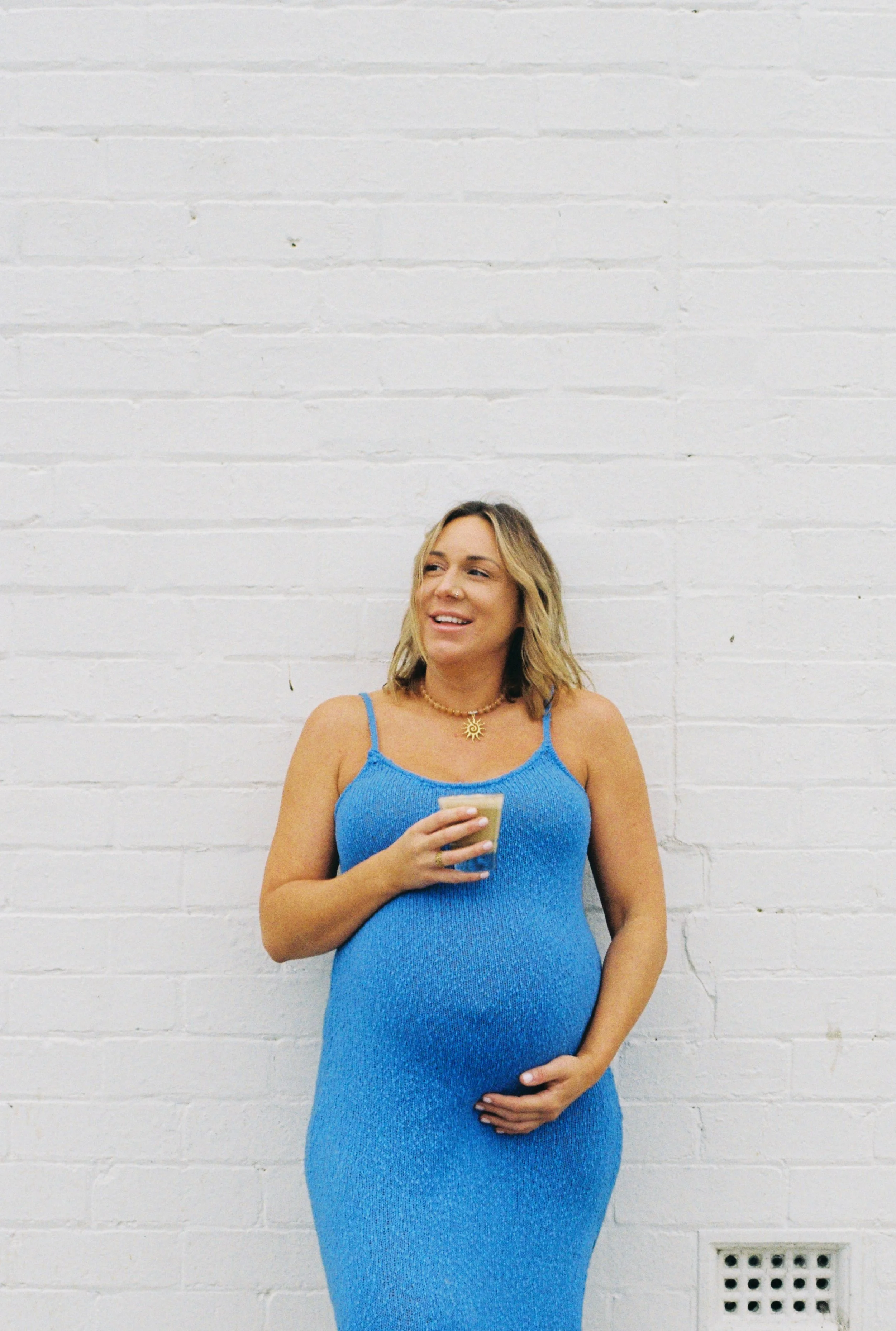 A woman wearing a blue dress and a sun-shaped necklace, holding a drink, standing against a white brick wall, looking to the side and smiling.