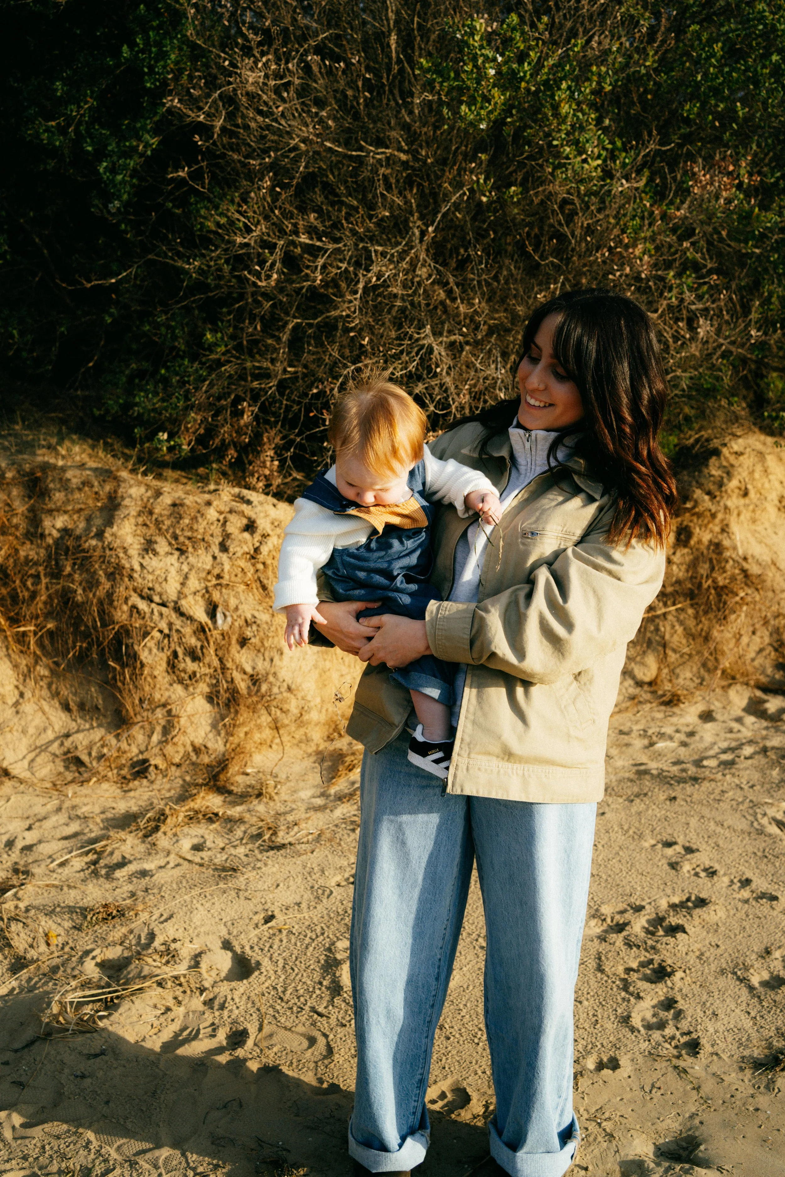 Woman wearing light-colored jacket and jeans holding a small child with blonde hair on a sandy beach with a large bush or tree in the background.