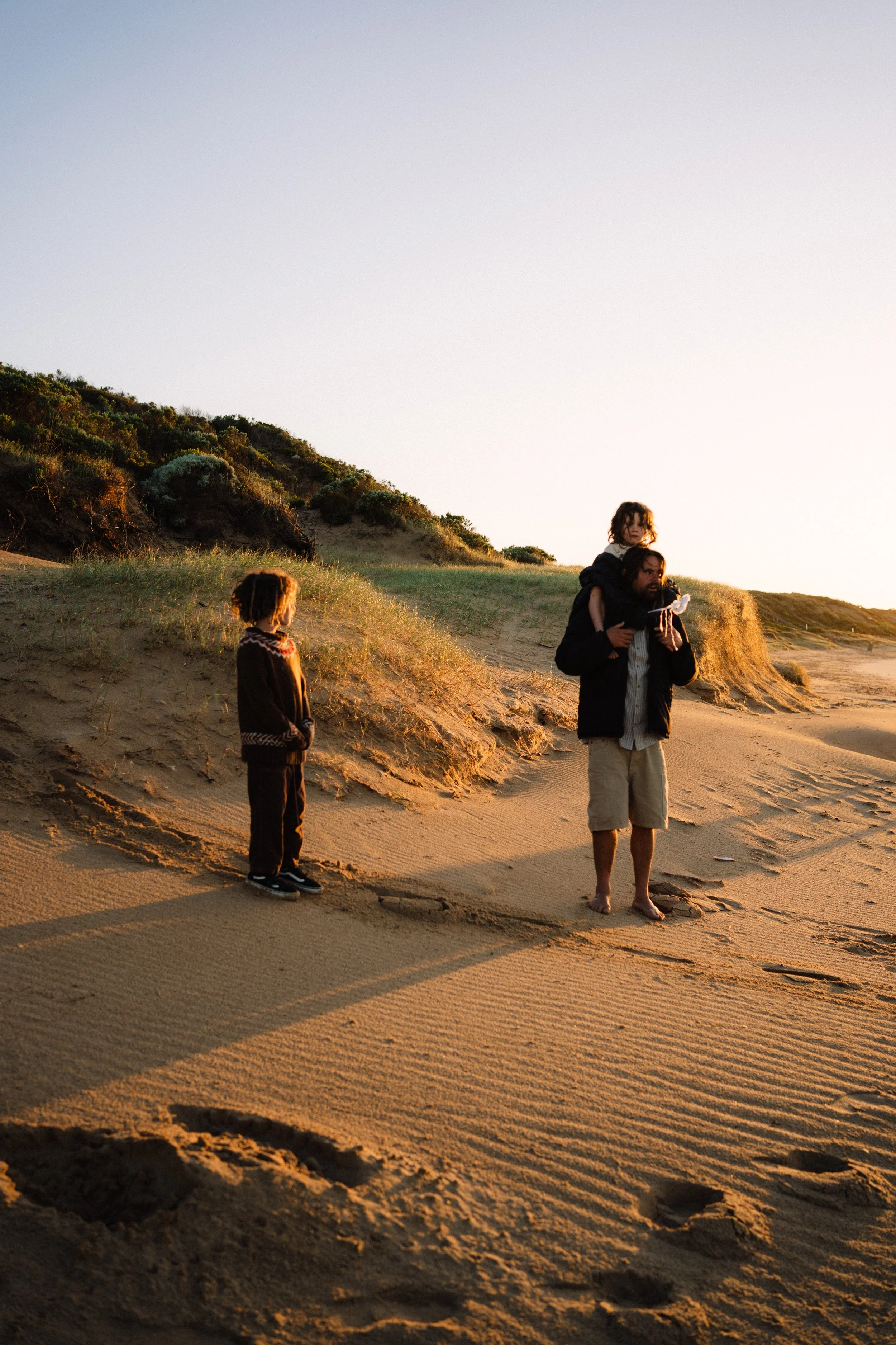 A man carrying a girl on his shoulders on a sandy beach during sunset, with a boy standing nearby and green hills in the background.