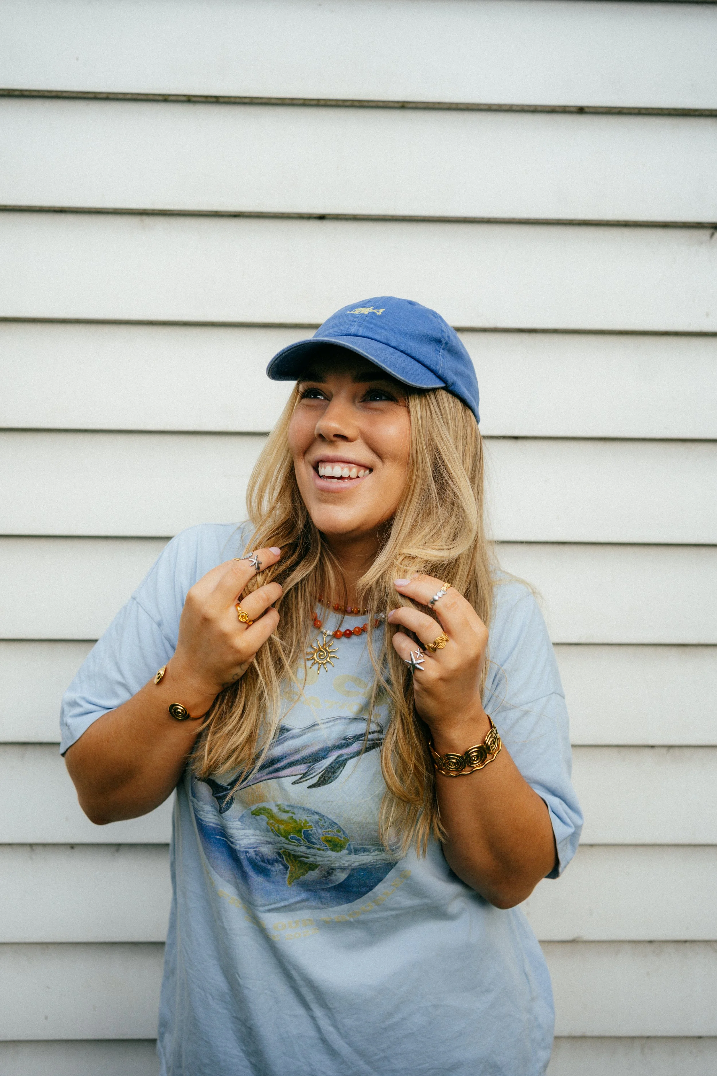 A young woman with long blond hair, wearing a blue cap and a light blue T-shirt with a globe graphic, smiling and looking up, standing against a white wooden wall, adorned with multiple rings, bracelets, and necklaces.