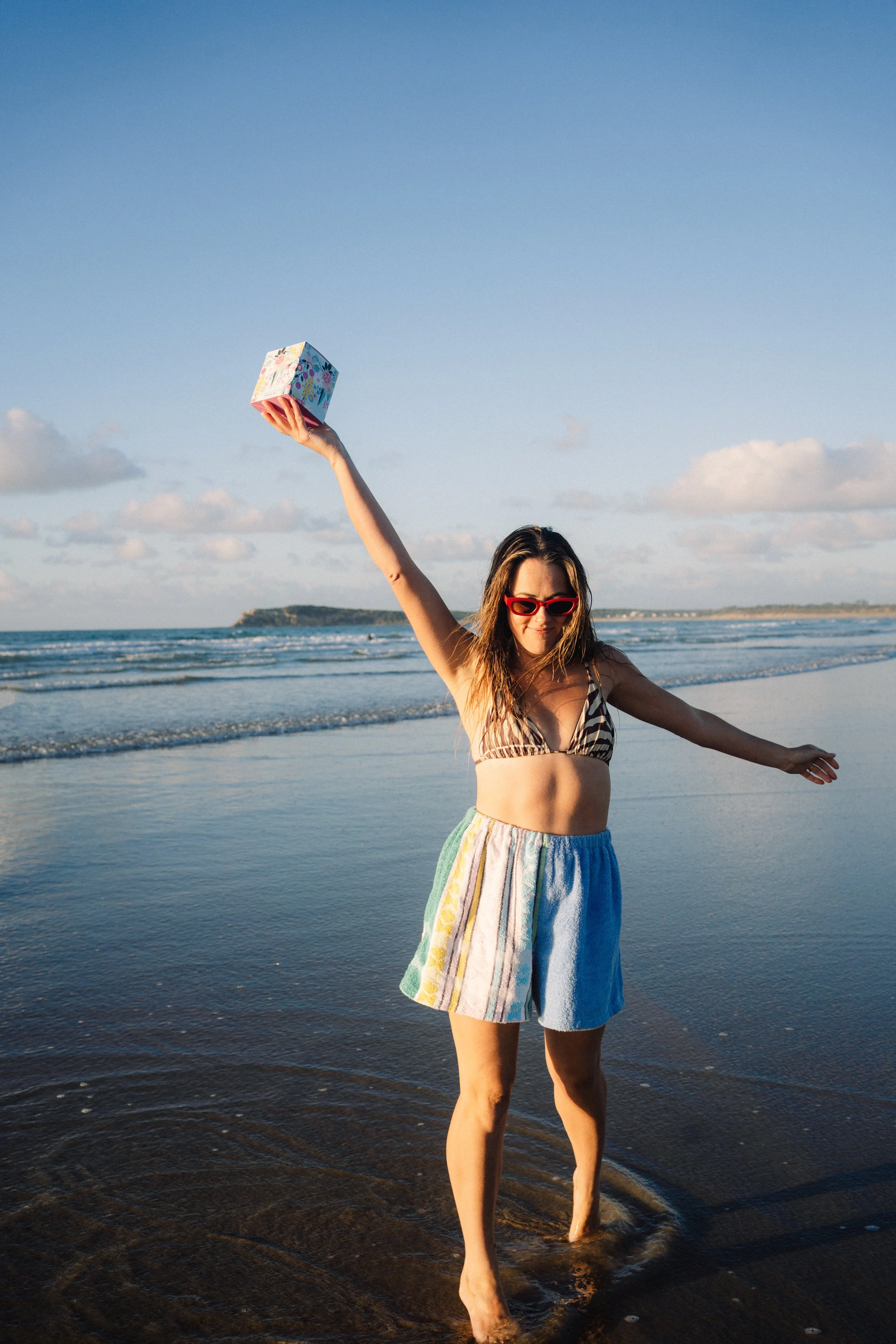 A woman with long hair, wearing red sunglasses, a striped bikini top, and a colorful striped towel skirt, is standing in shallow ocean water during sunset, holding a decorated box high above her head.