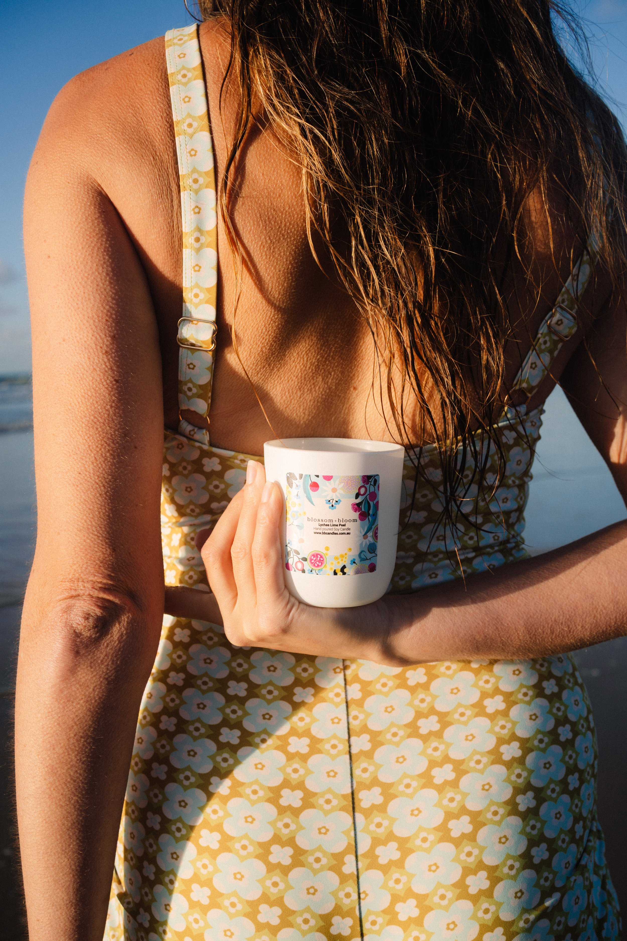Close-up of a woman in a yellow dress with white flowers, holding a white mug with a floral design, standing outdoors in bright sunlight.