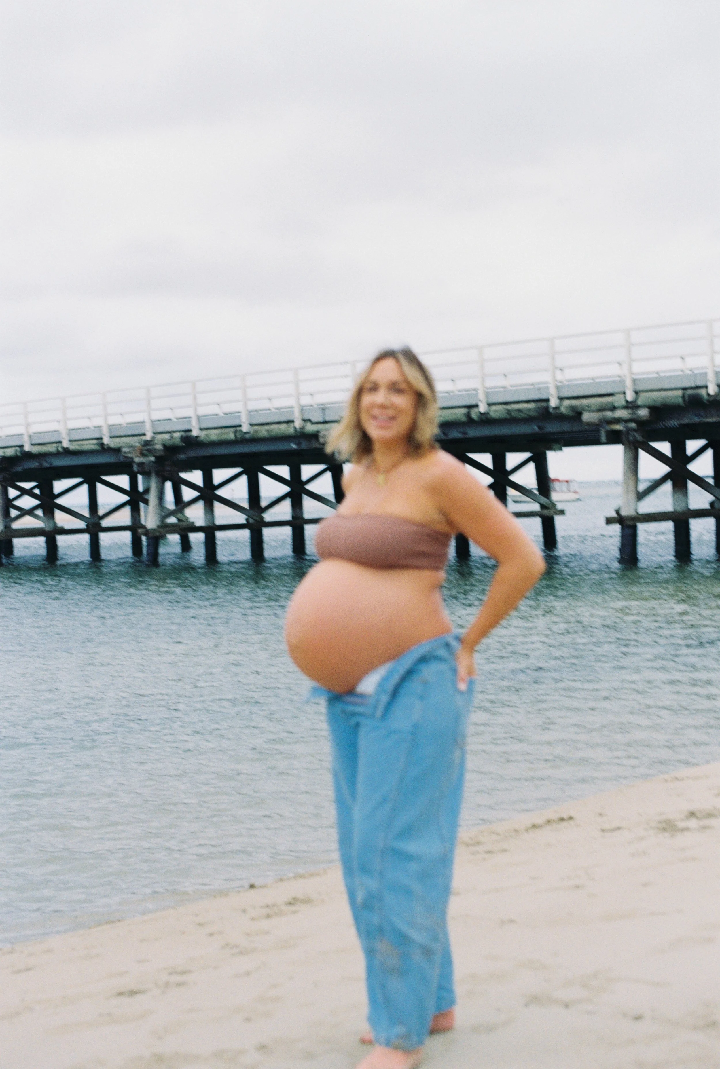 A pregnant woman with blonde hair wearing a brown tube top and blue jeans standing on a beach near water with a pier in the background.