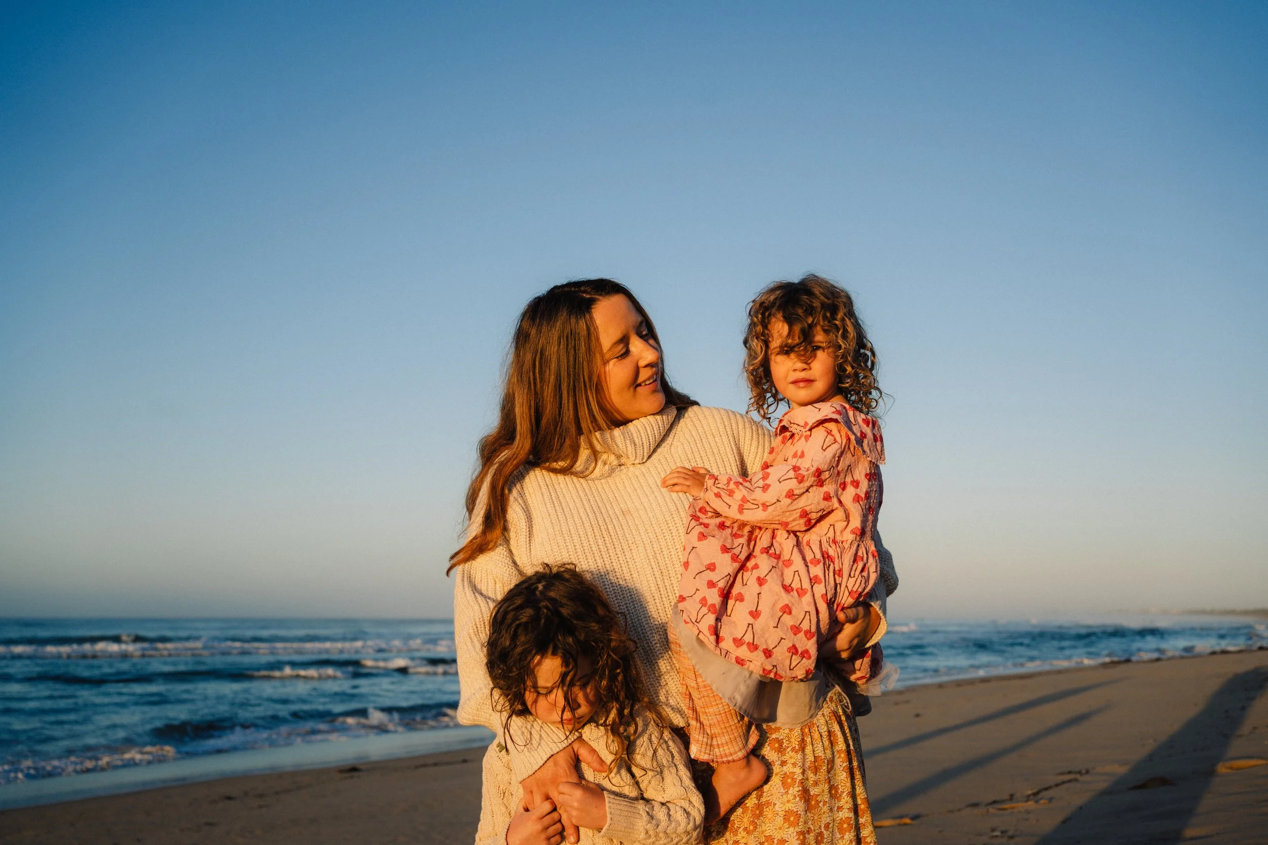 A woman holding a young girl on a beach at sunset, with two children partially visible in front, the ocean and sky in the background.