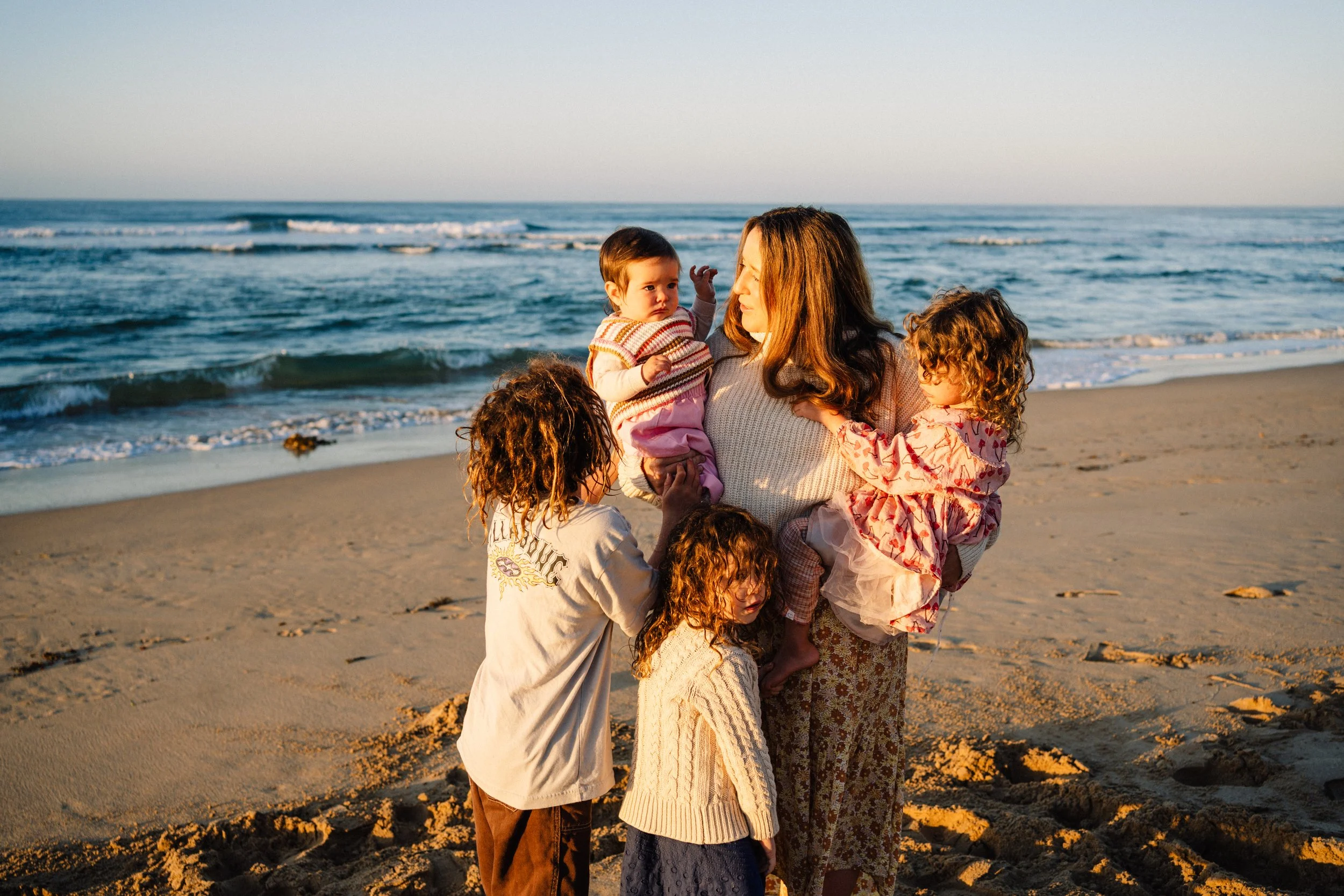 A woman holding a baby while surrounded by children on a sandy beach during sunset, with the ocean waves in the background.