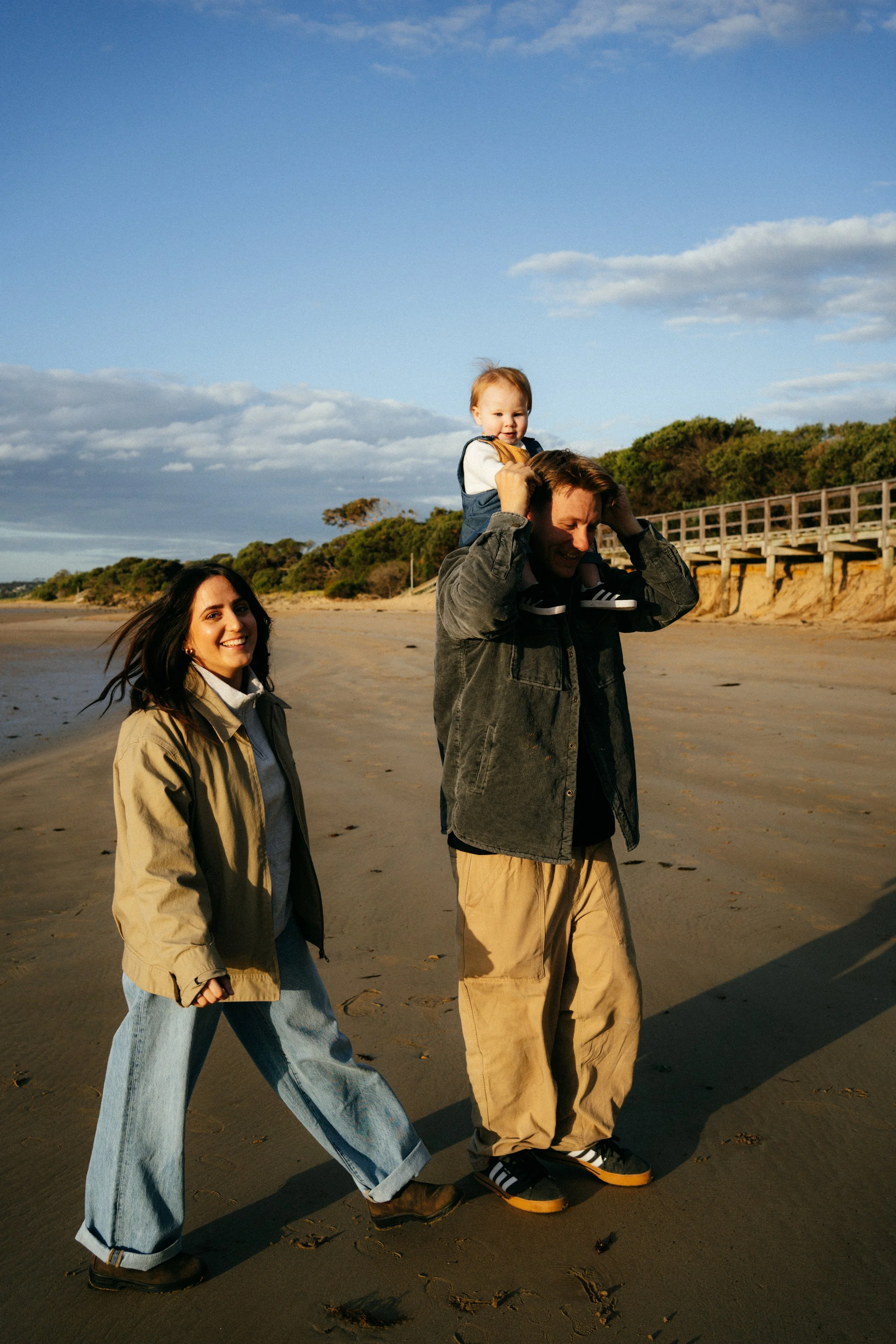 A group of three people walking on a sandy beach with dry footprints, a wooden walkway, trees, and partly cloudy sky in the background, enjoying a sunny day.