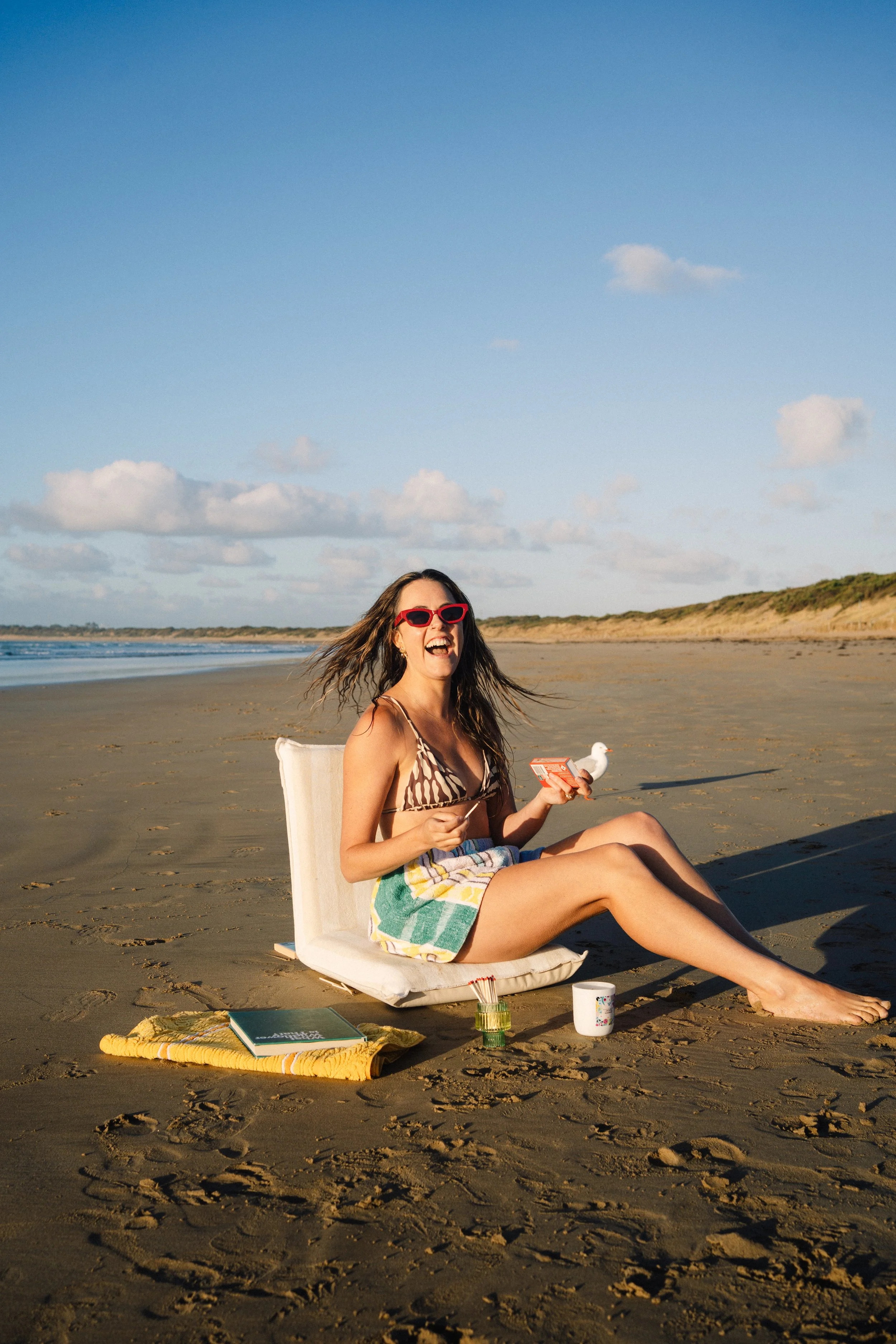 Woman with long hair and red sunglasses, sitting on a beach chair on the sand, smiling and laughing, holding a snack, with notebooks, cups, a towel, and a small container nearby, under a partly cloudy sky.