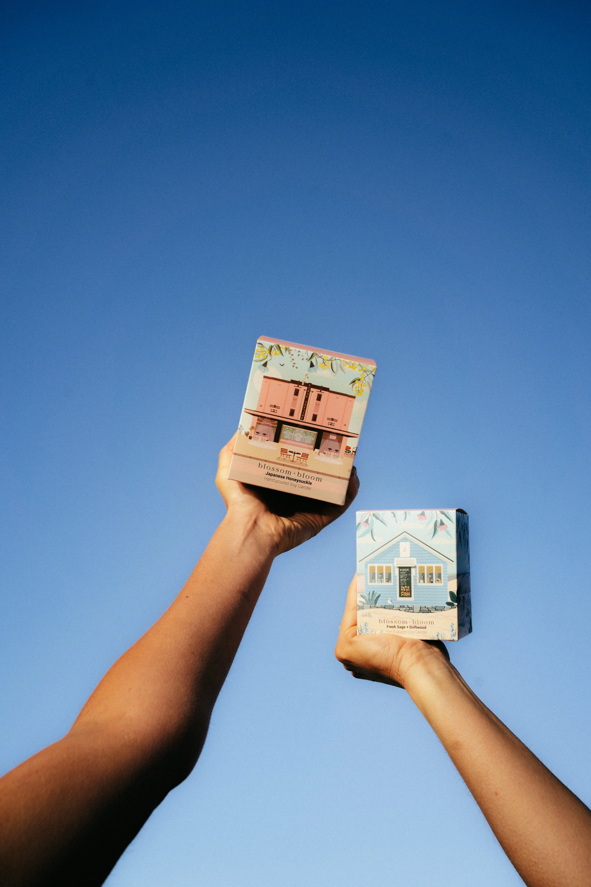 Two hands holding boxes of Blossom Bloom candles against a clear blue sky.