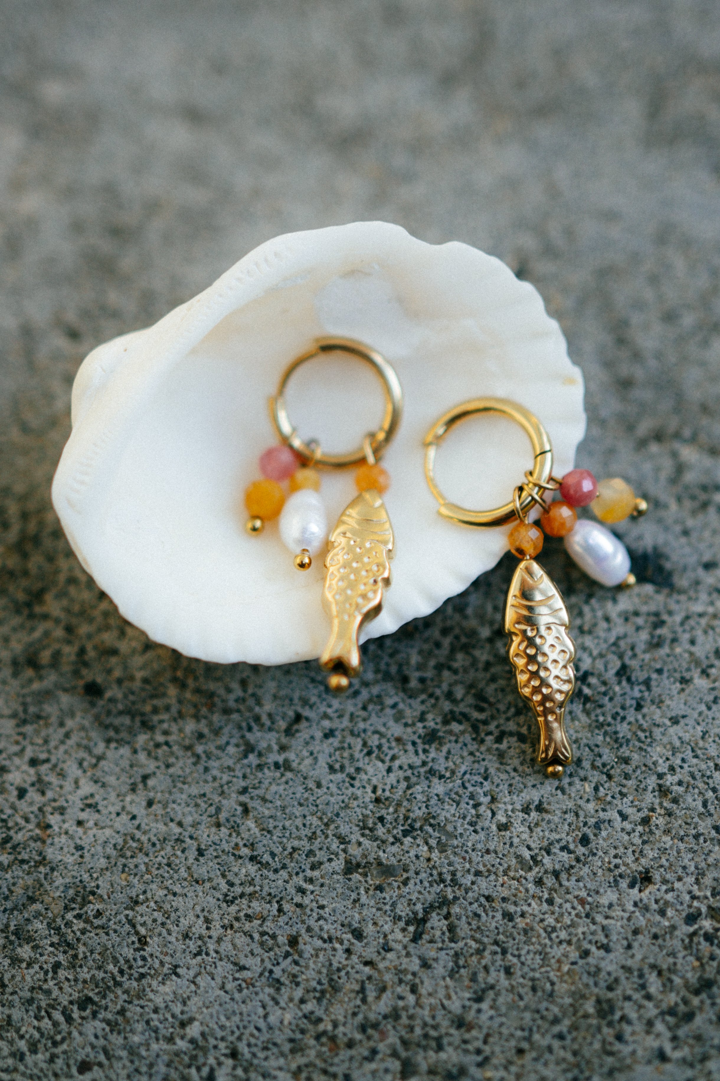Gold hoop earrings with colorful beads and fish-shaped charms, placed on a white seashell on a sandy surface.