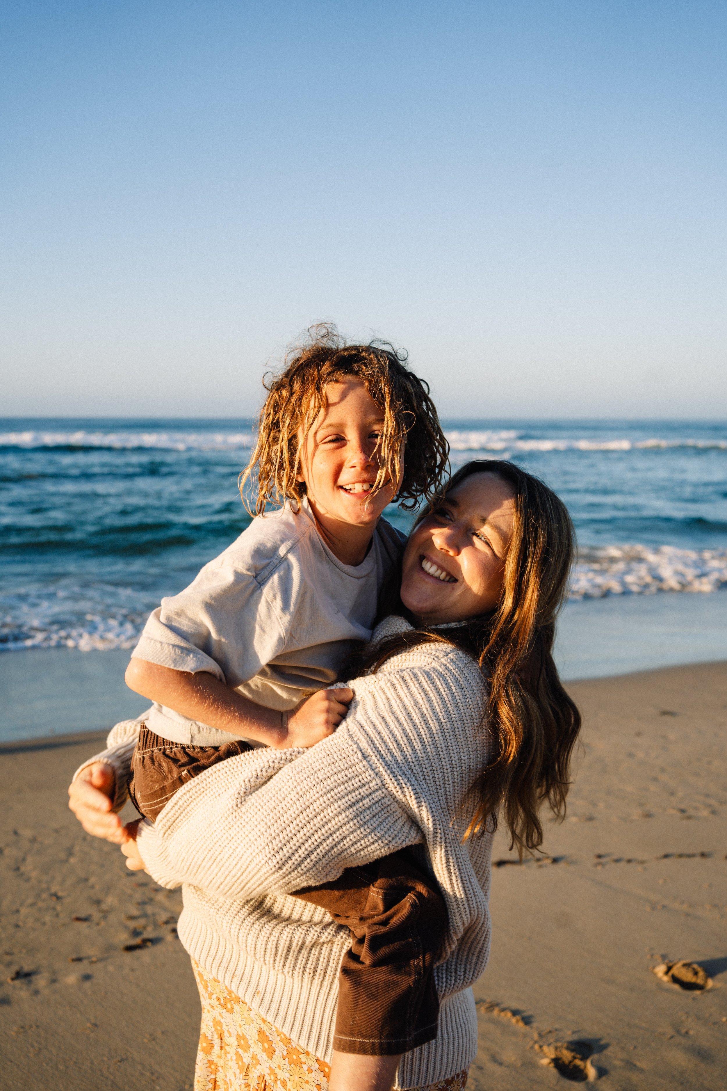 A woman holding a child on her shoulders at the beach, both smiling and enjoying the sunset