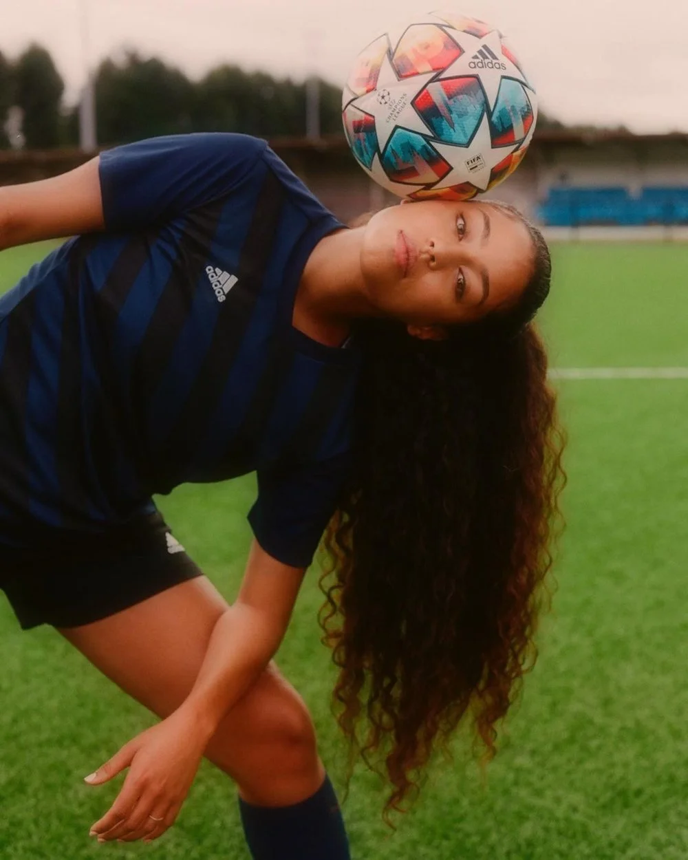Female soccer player with long curly hair in a dark blue striped Adidas jersey on a soccer field, balancing a colorful Adidas UEFA Champions League soccer ball on her shoulder.
