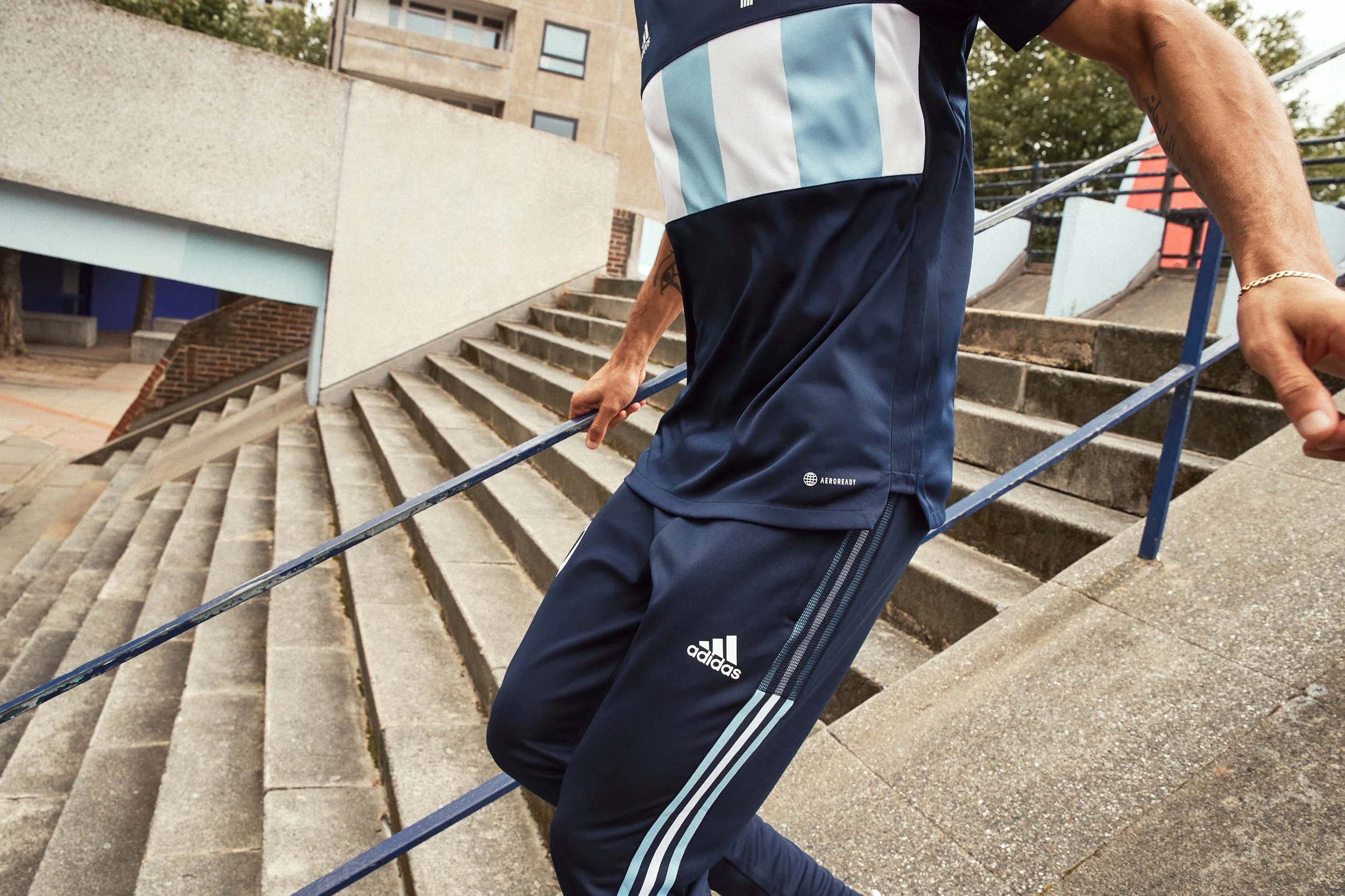 Close-up of a person descending outdoor concrete stairs while holding a blue handrail, wearing navy blue adidas athletic pants and a matching shirt with light blue and white stripes.