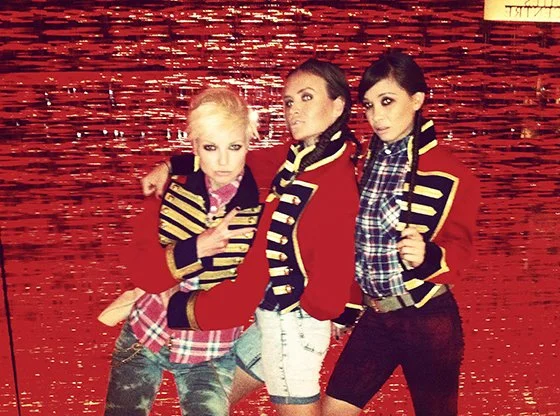 Three young women posing in front of a red water background, wearing matching striped jackets with gold trim.