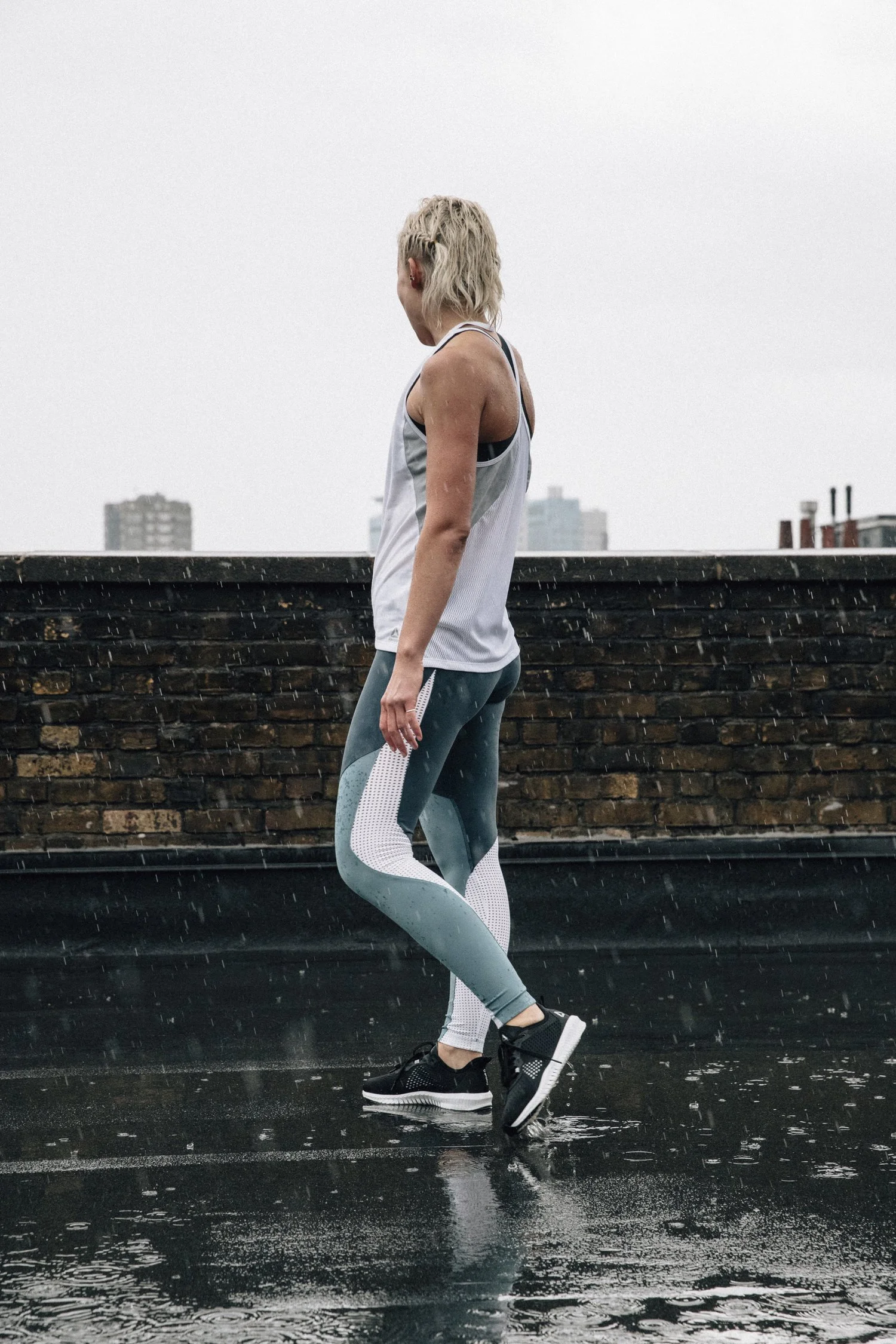 A woman in athletic wear walking on a wet rooftop on a rainy day, with city buildings in the background.