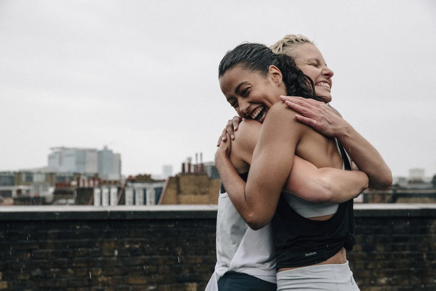 Two women embracing and smiling on a rooftop, with a city skyline in the background on a cloudy day.