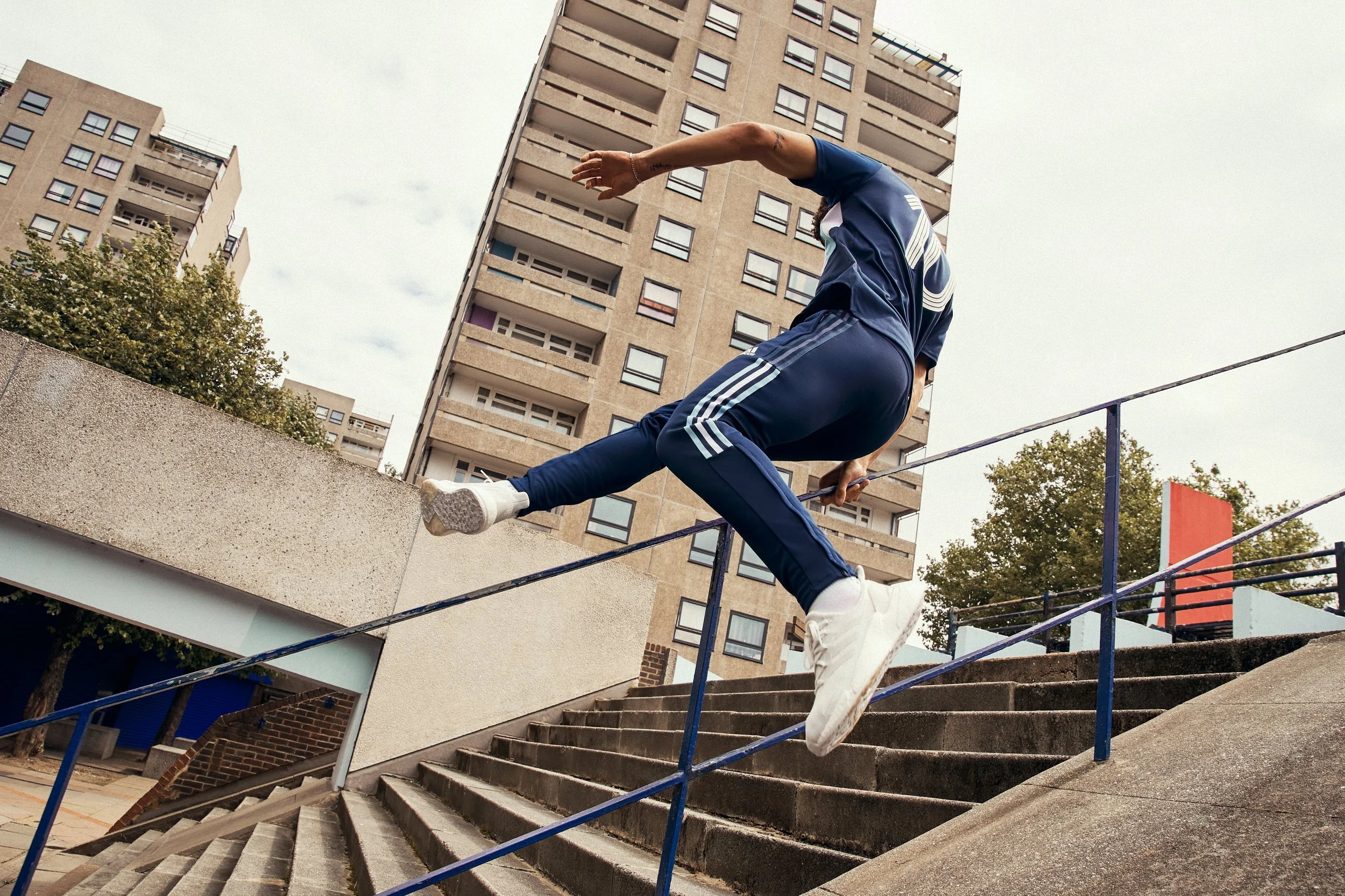 A person in athletic clothing running and jumping over a handrail on outdoor stairs in an urban setting with tall apartment buildings in the background.
