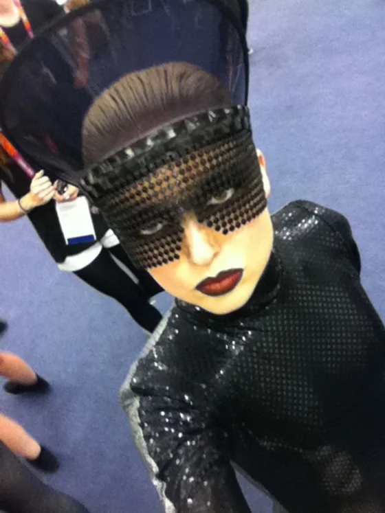 Person with elaborate makeup and headpiece wearing a black, shiny, textured outfit at an indoor event.