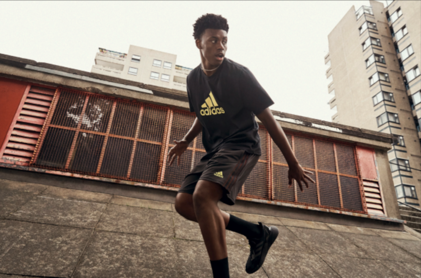 Young man with dark skin and curly hair playing soccer outdoors in urban environment, wearing black Adidas sportswear, on concrete surface, with tall apartment buildings in the background.