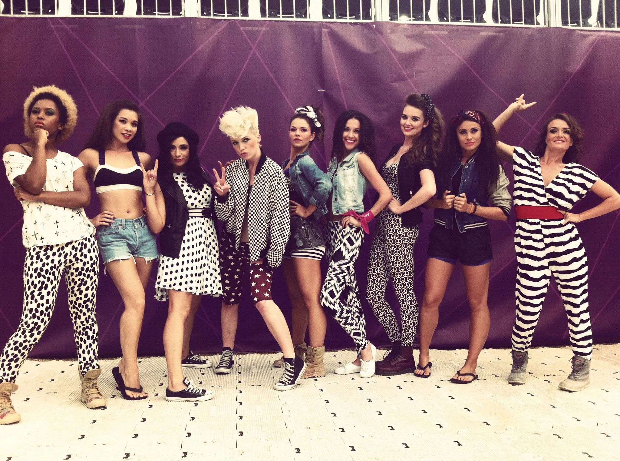 A group of ten women standing in a line in front of a purple backdrop, wearing casual and stylish clothing with various patterns like polka dots, stripes, and geometric prints, posing playfully for the photo.