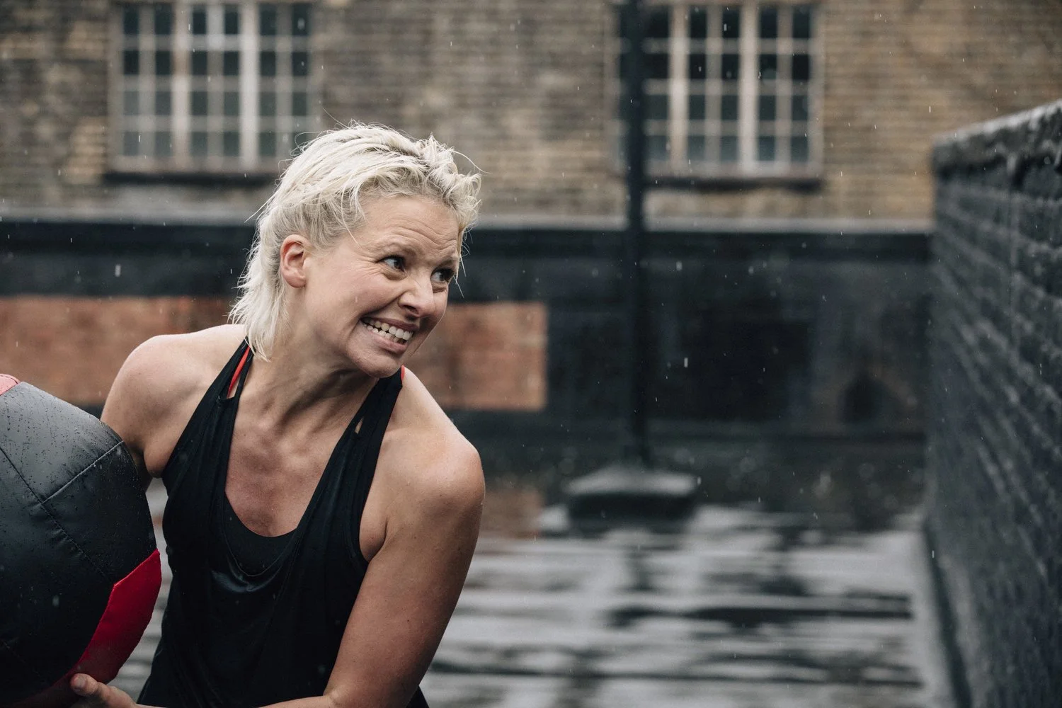 A woman with short blonde hair smiling while holding a medicine ball outdoors in rainy weather.