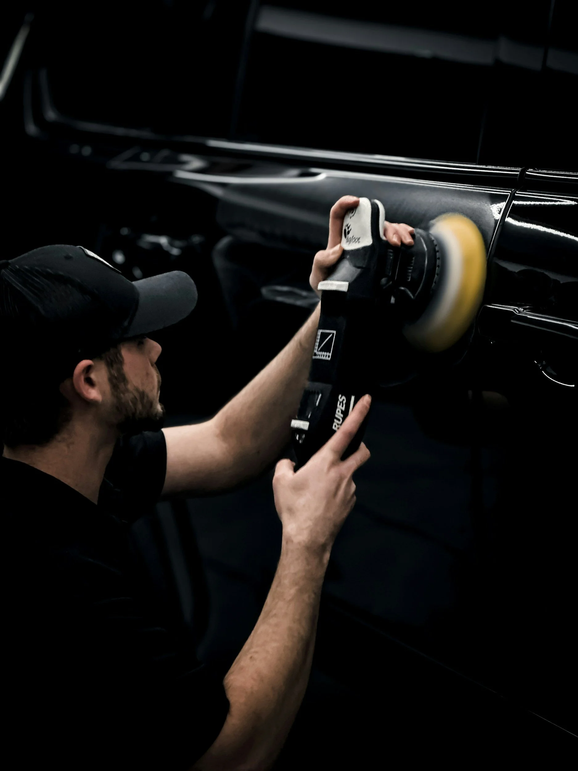 A man wearing a black cap polishing a black car with a yellow buffer.