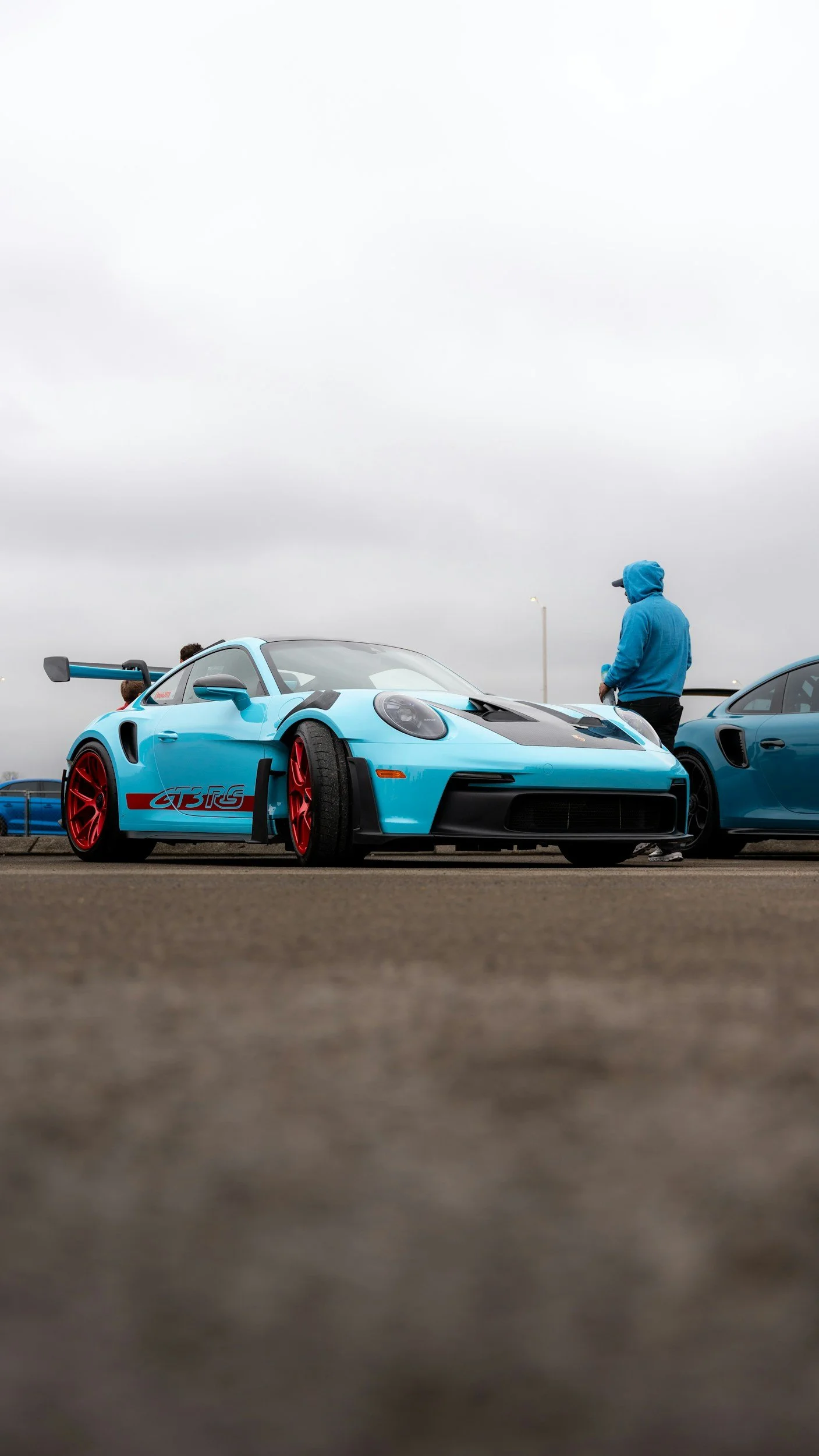 A blue race car with red wheels parked on an overcast day, with a person in a blue hoodie standing nearby.