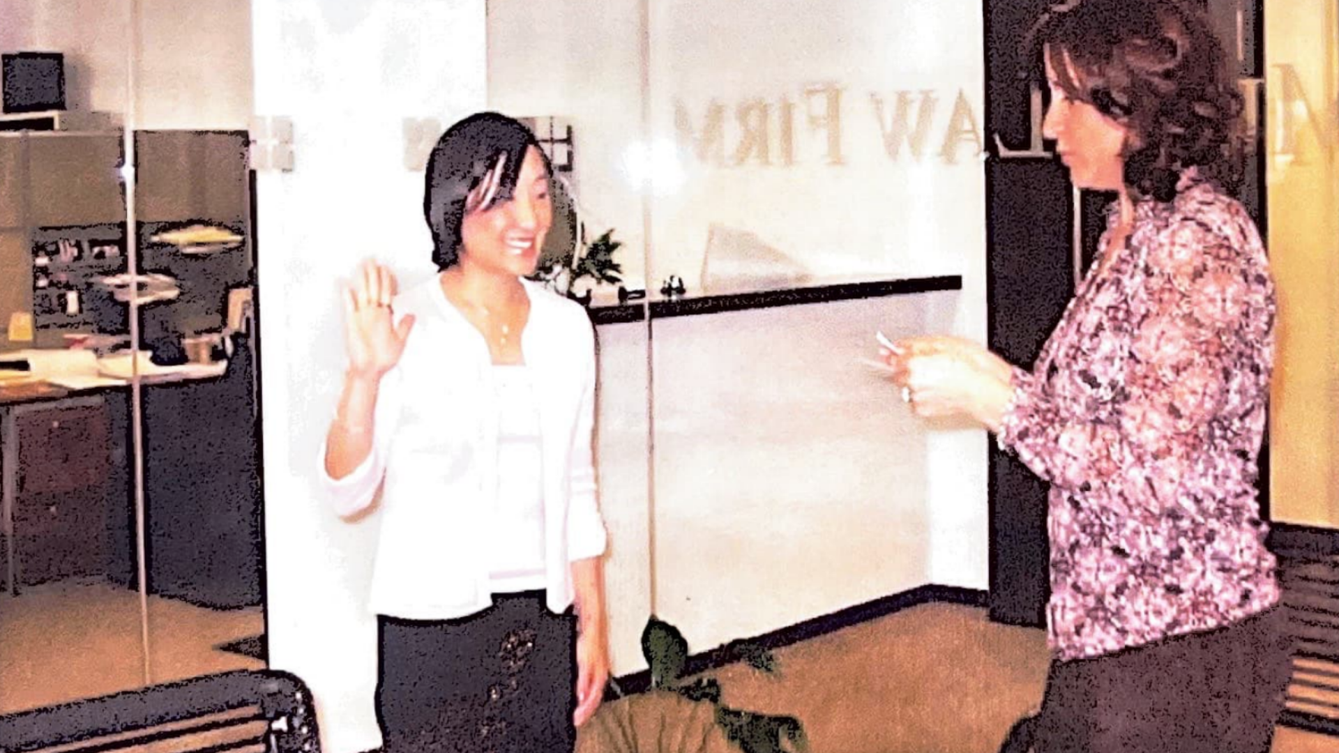 Two women standing in an office setting, one smiling and holding her right hand up as she is being sworn in as an attorney while the other looks down at something in her hands.