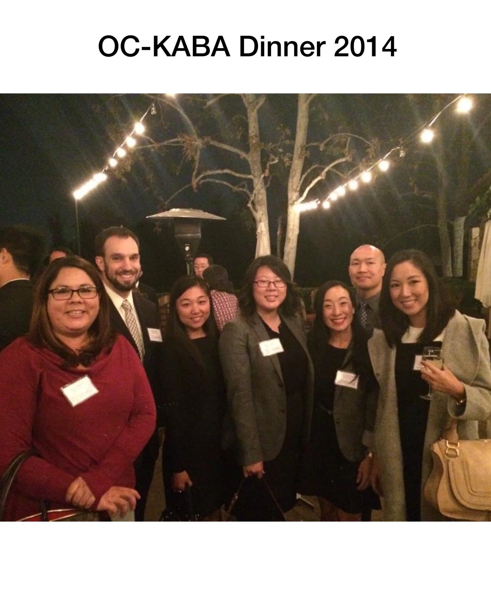 Group of people attending OC-KABA Dinner in 2014 at an outdoor event during the evening, with string lights and a tree in the background.