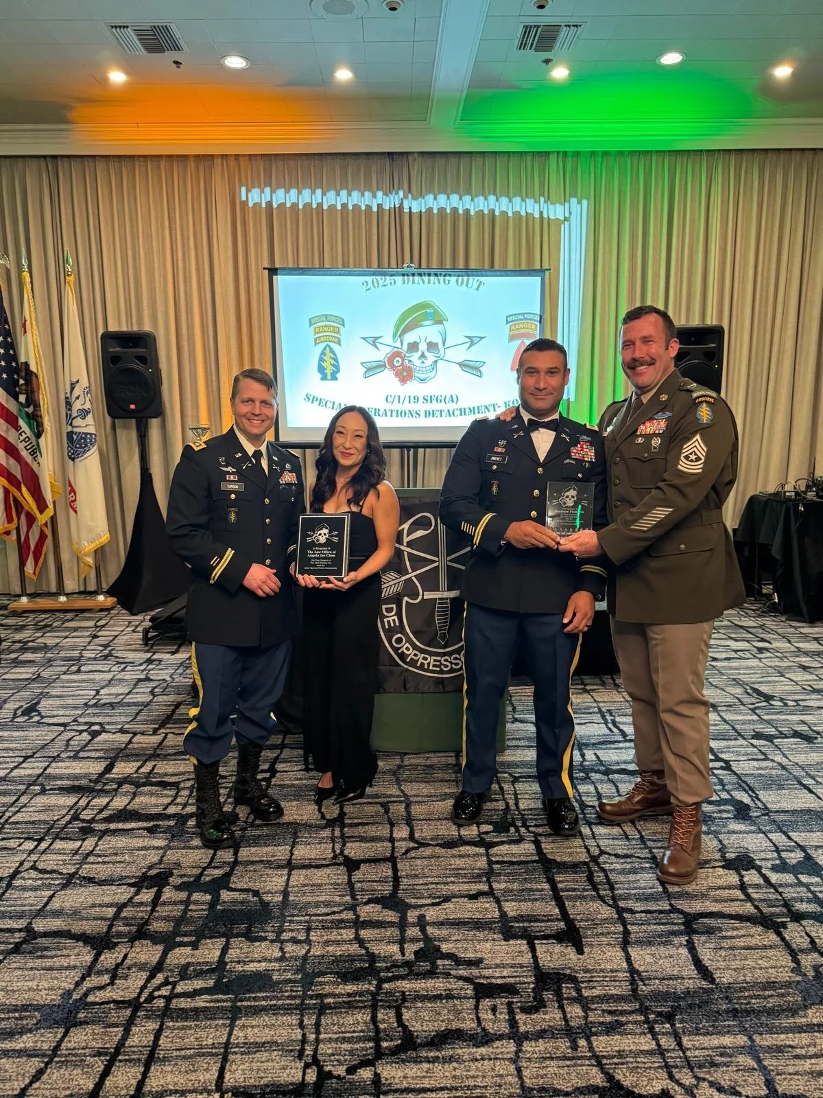 Four people, three men in military uniform and one woman dressed in all black, pose for a photo in an award ceremony. Three of them are holding an award.