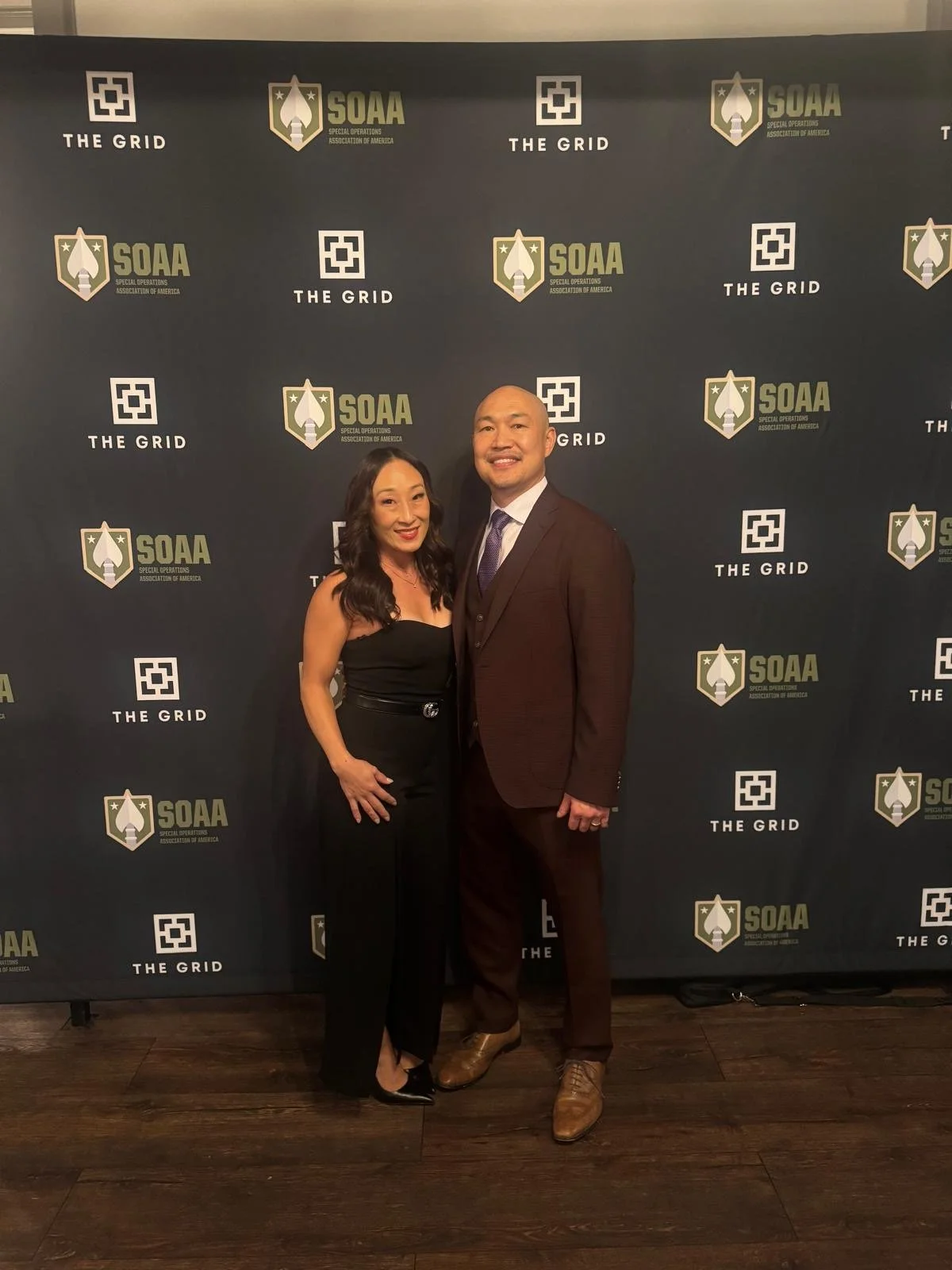 A woman in a black one piece and a man in a brown suit stand next together with a smile. The back drop has logos of different organizations and companies.