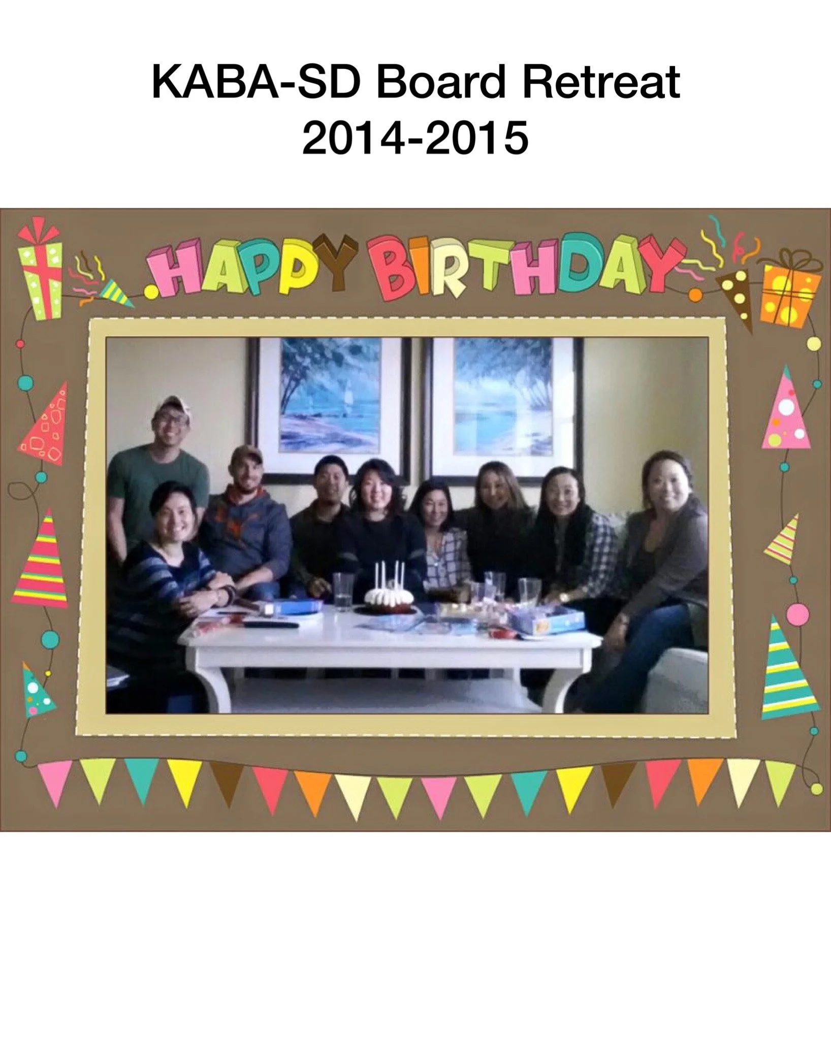 A group of nine people sitting around a table during a birthday celebration, with a birthday cake and party decorations, inside a room with framed artworks on the wall.