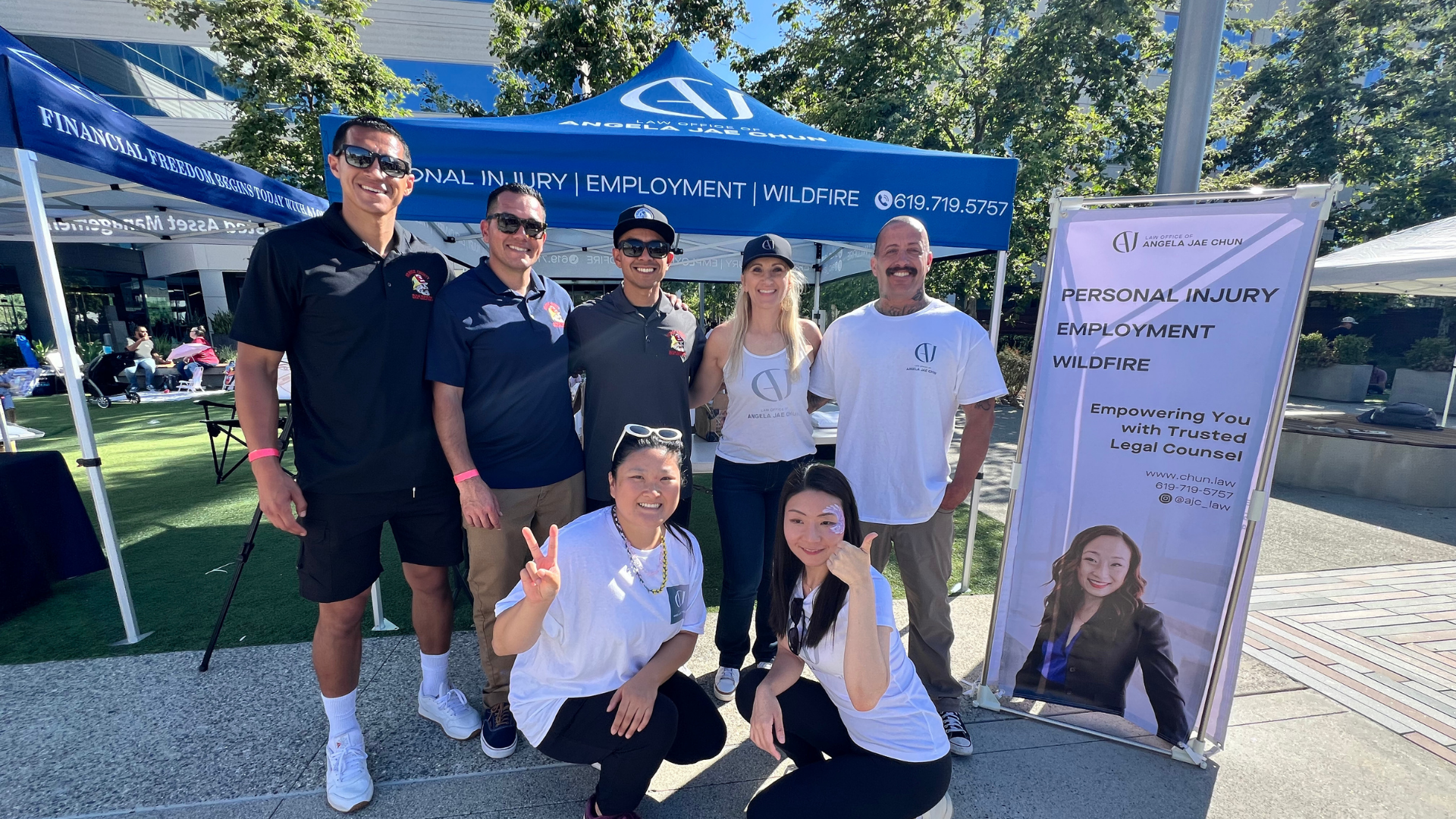 Group of people at an outdoor event standing in front of a blue tent and a white banner. The banner displays the logo and contact information for the Law Office of Angela Jae Chun, focusing on personal injury, employment, and wildfire legal services. The people are smiling and posing for the photo, some wearing matching shirts with the law office logo.