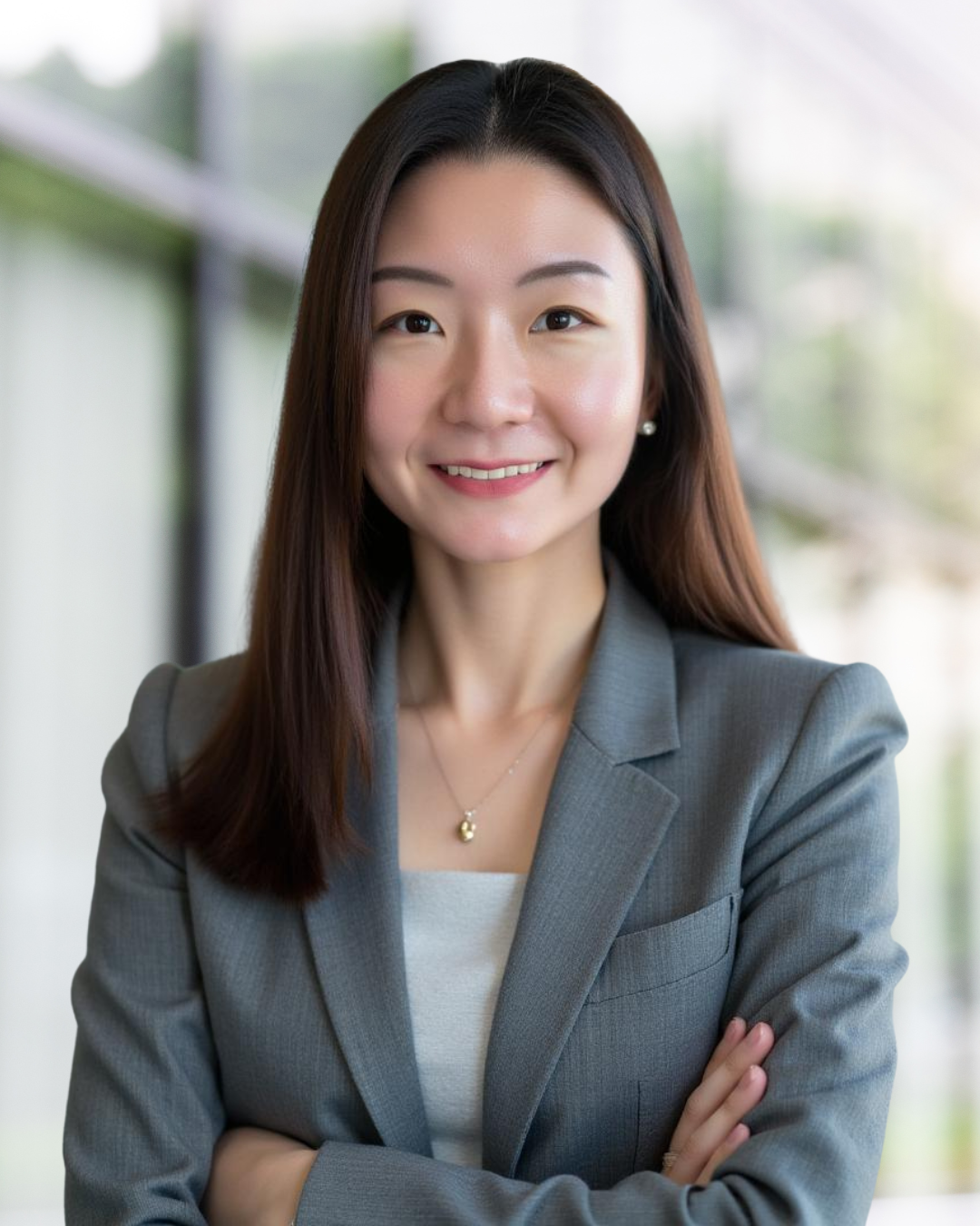 A young woman with long dark hair, wearing a black blazer and black top, looking at the camera with a neutral expression, indoors with blurred warm lighting and large windows in the background.