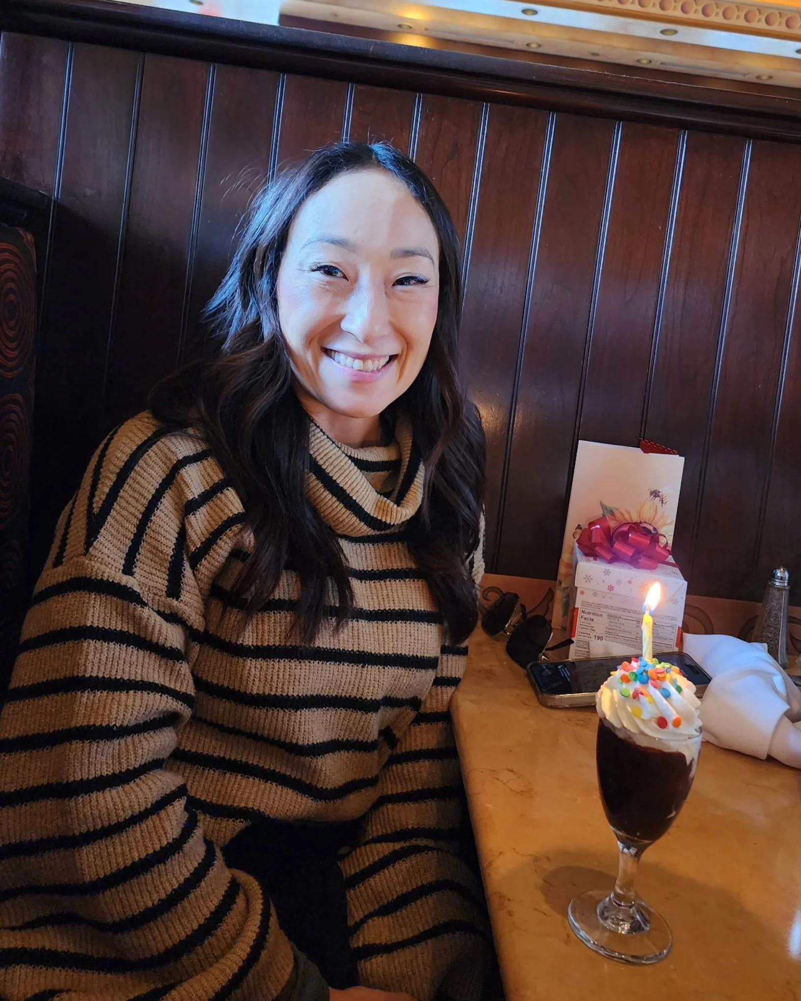 Woman smiling while seated at a restaurant table with a dessert topped with a lit candle.