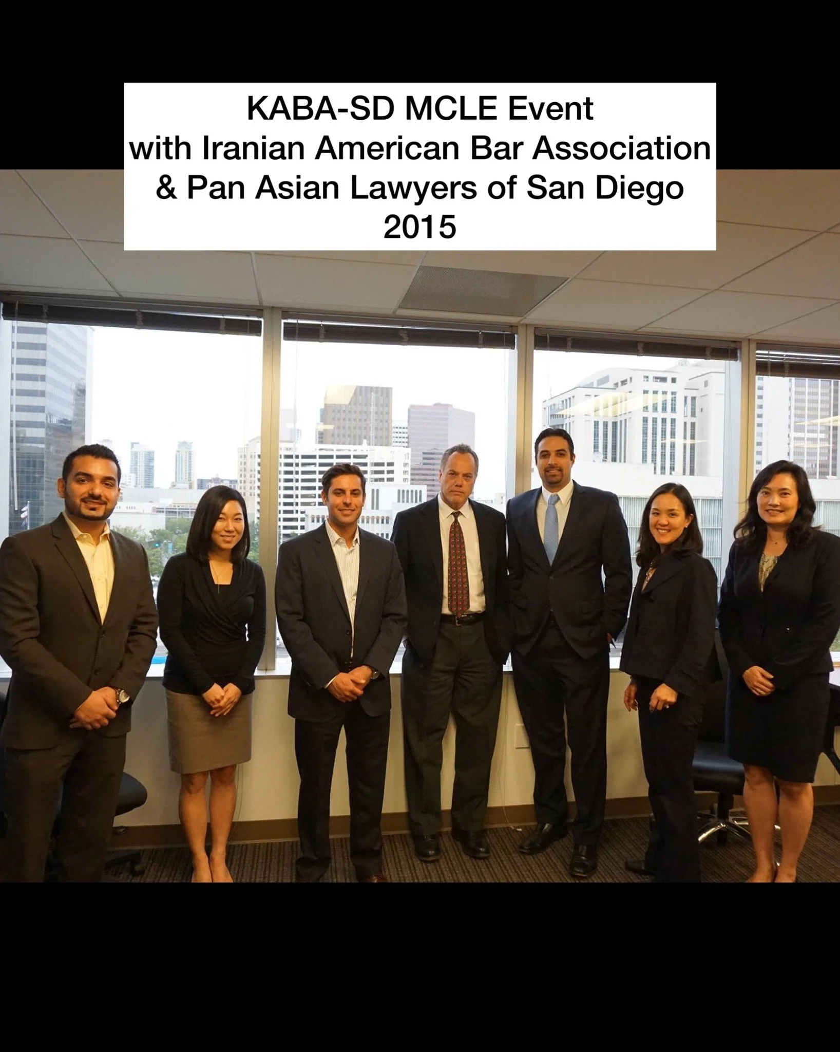 Group of seven professionals in business attire standing in office with large windows and cityscape background