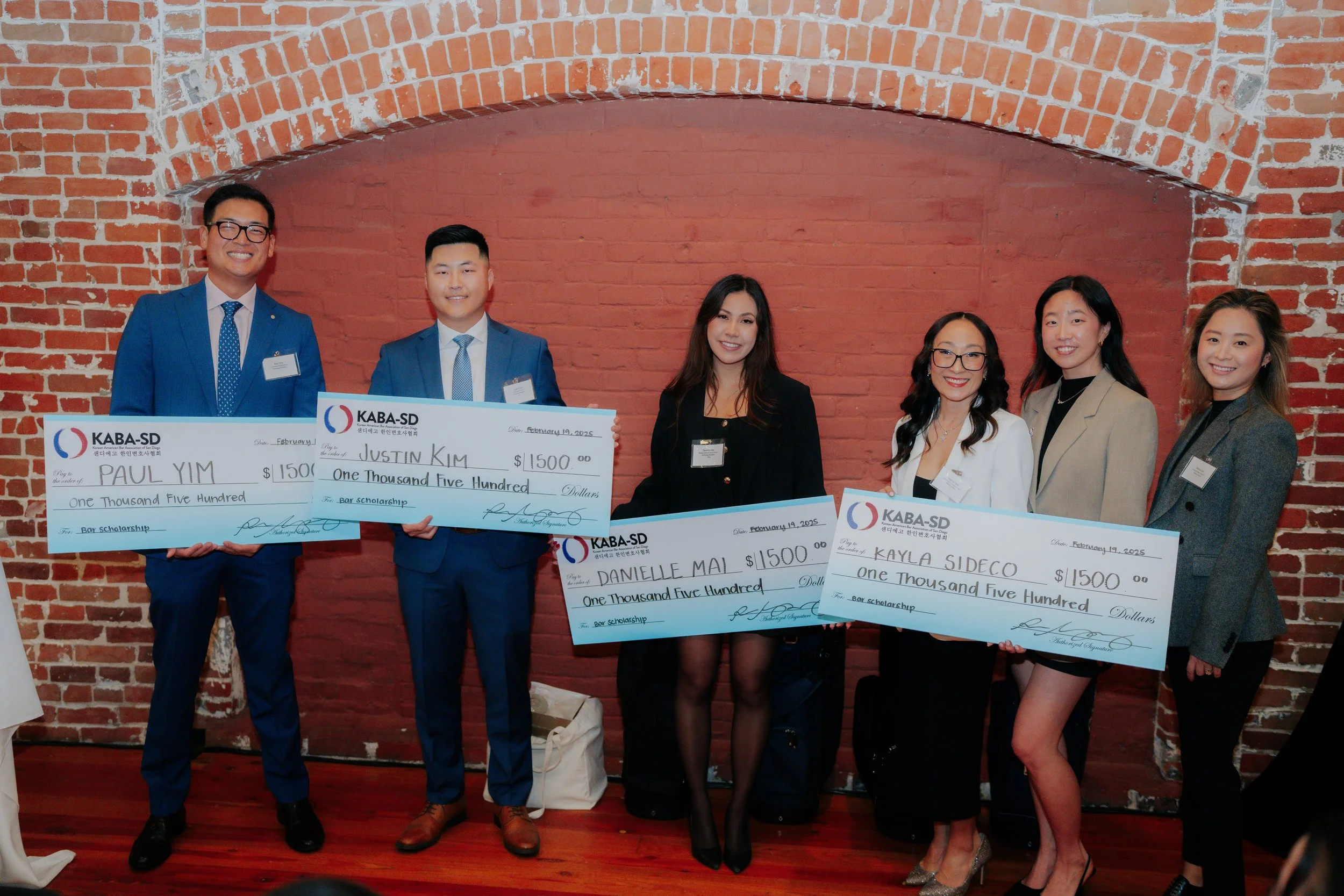 Group of six young adults standing against brick wall, holding large checks for $1500 each, celebrating KABA-SD scholarship awards at an indoor ceremony.