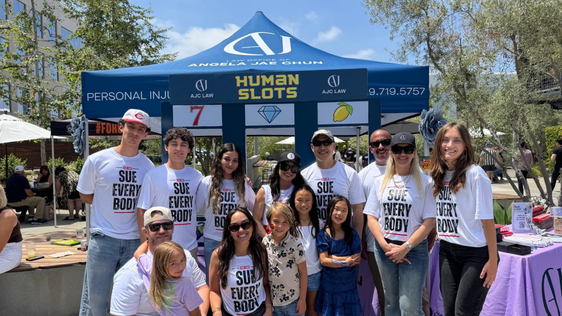 Staff of the Law Office of Angela Jae Chun, wearing T-shirts that say "Sue Everybody" while smiling and posing for a photo in front of a tent at an outdoor event for the OC Bomberos Cornhole Tournament.