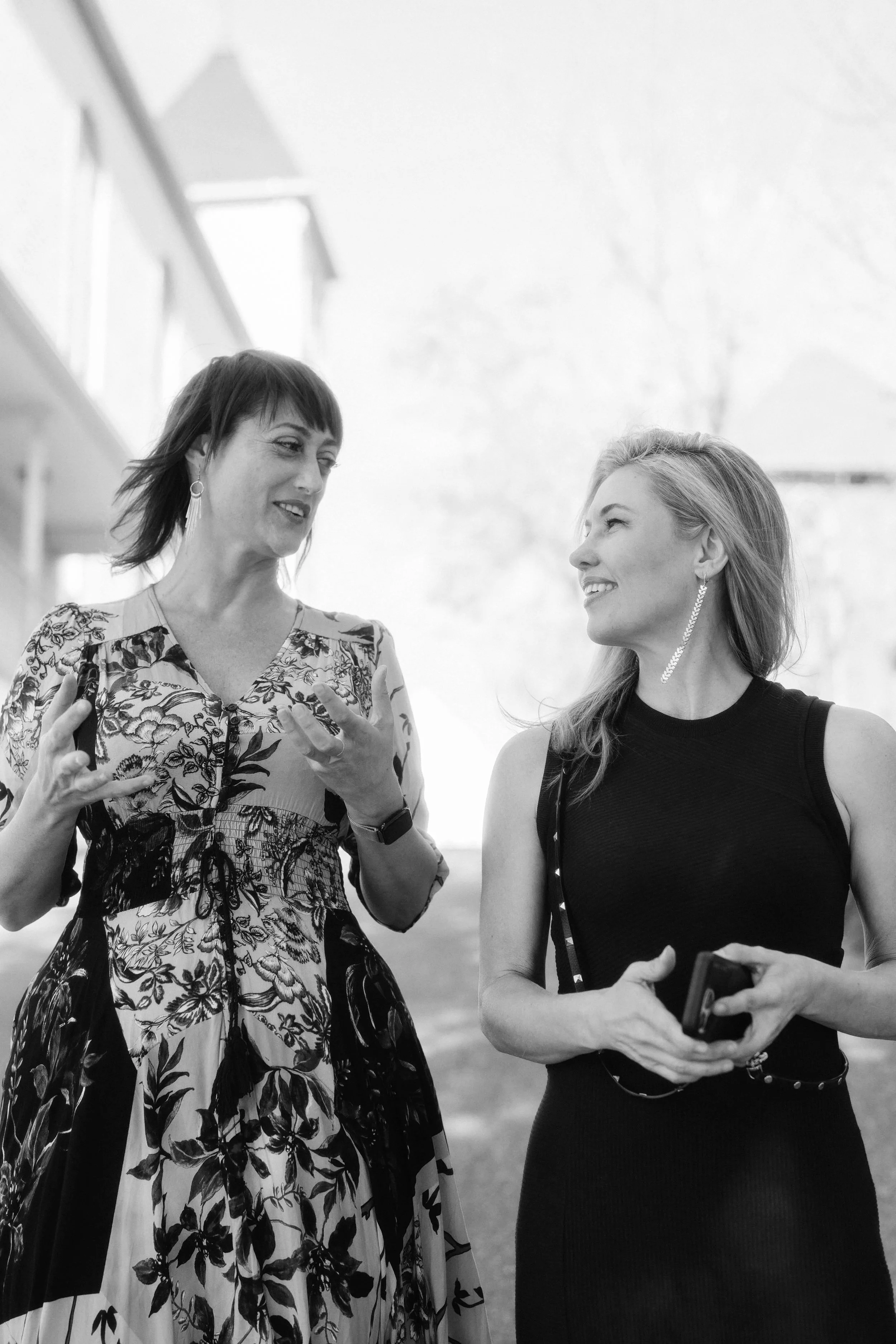 Two women are having a conversation outdoors. The woman on the left is wearing a floral dress, earrings, and a watch, and gesturing with her hands while speaking. The woman on the right is in a sleeveless black top, has long hair, earrings, and is holding a smartphone and a purse, smiling and listening attentively.