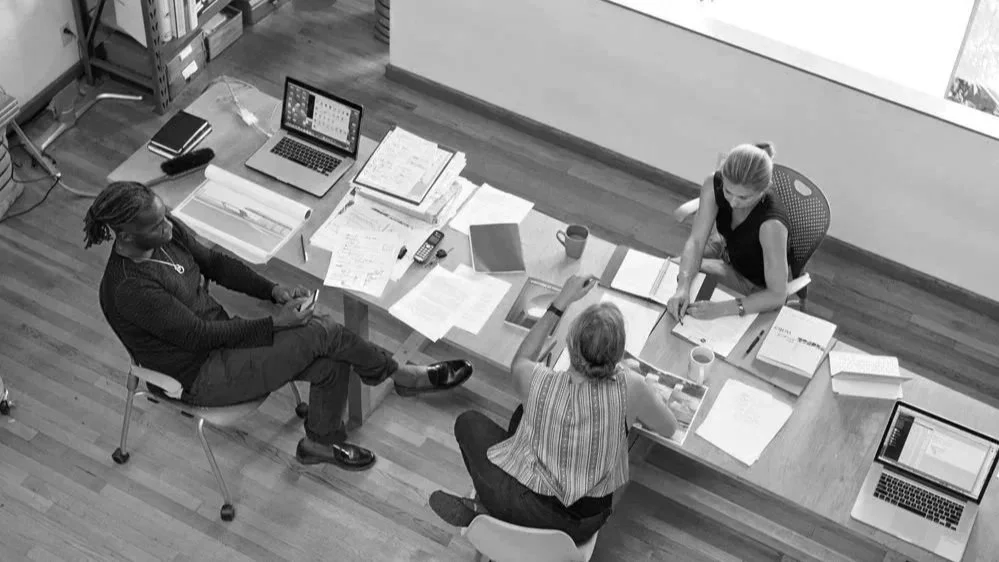 Three people sitting around a desk in an office, working with notebooks, papers, and laptops.
