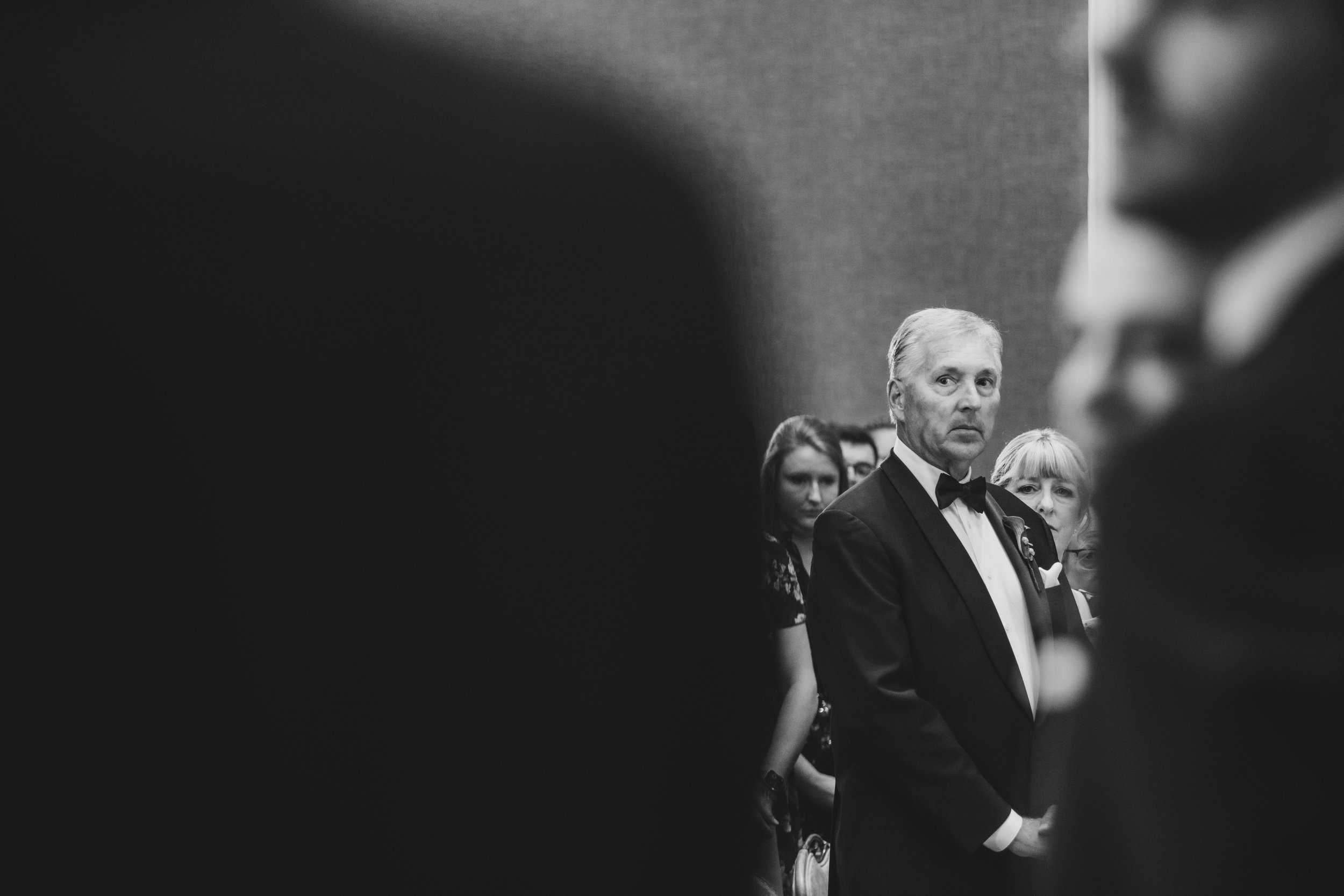 A black and white photo of formally dressed people, focusing on a man in a tuxedo with a bow tie, at an event or gathering.