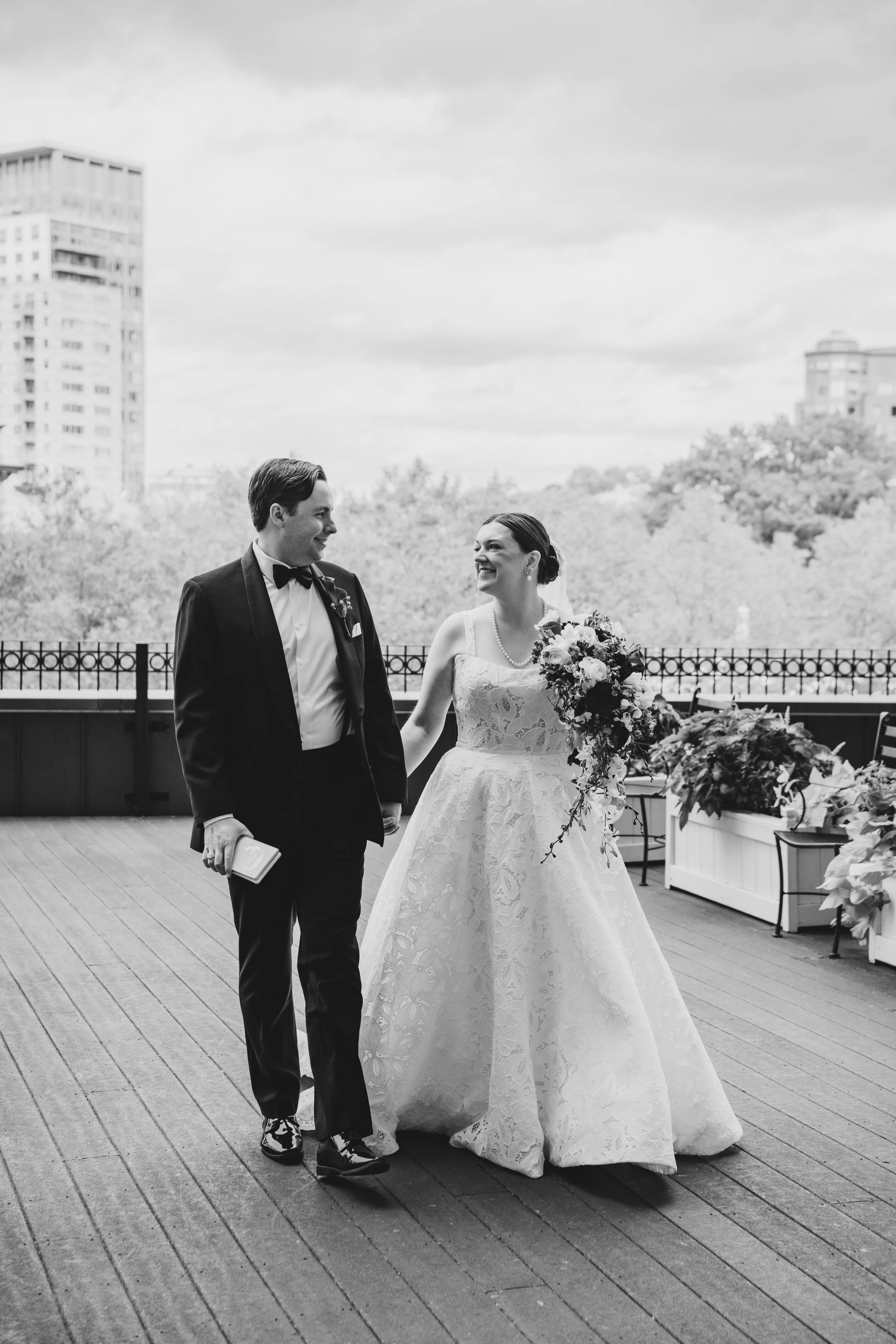 Black and white photo of a bride and groom holding hands on a wooden deck, smiling at each other, with city buildings and trees in the background.