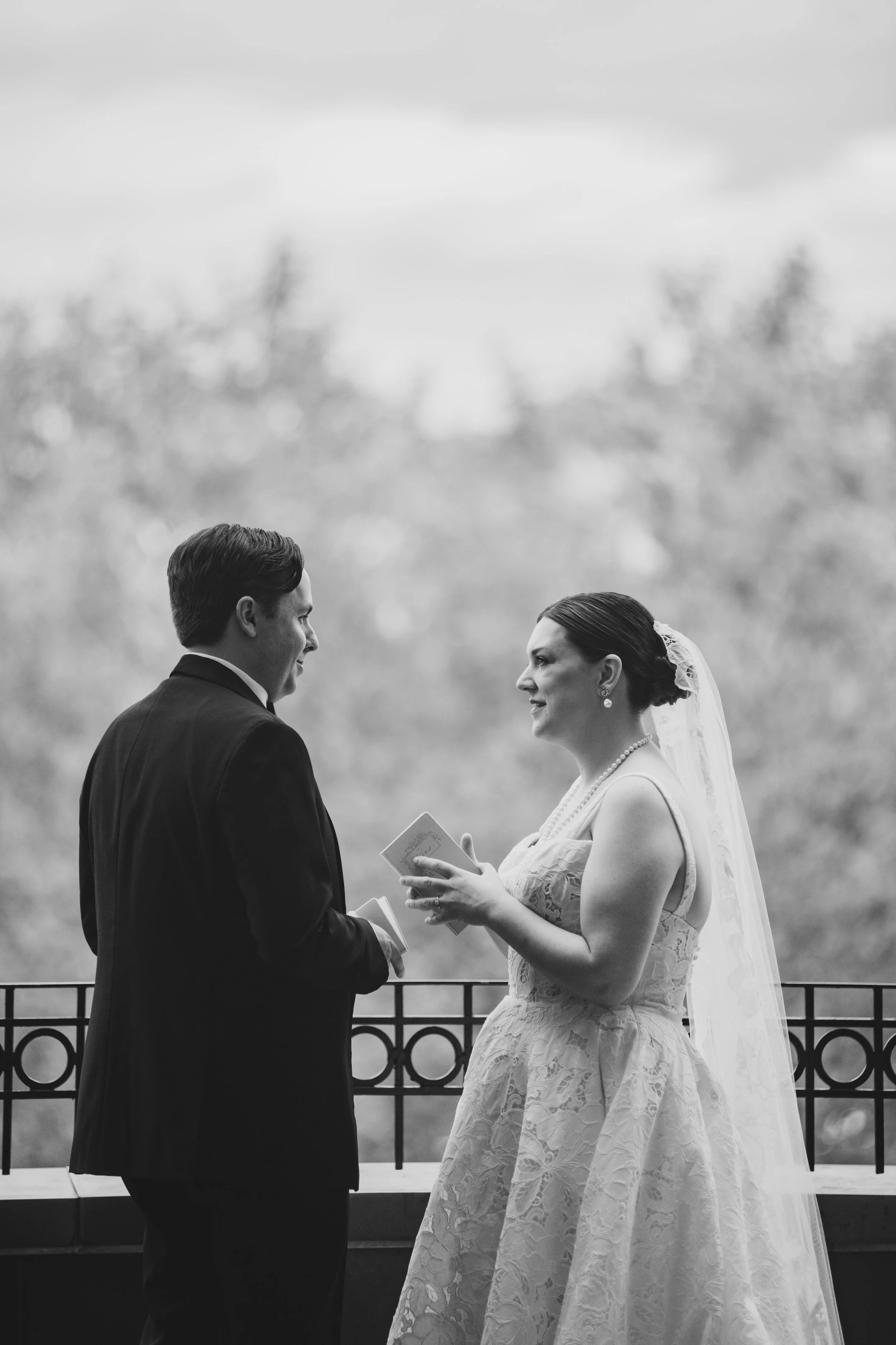 A bride and groom exchanging vows during their wedding ceremony outdoors, with the bride holding a small book or card, both smiling at each other.