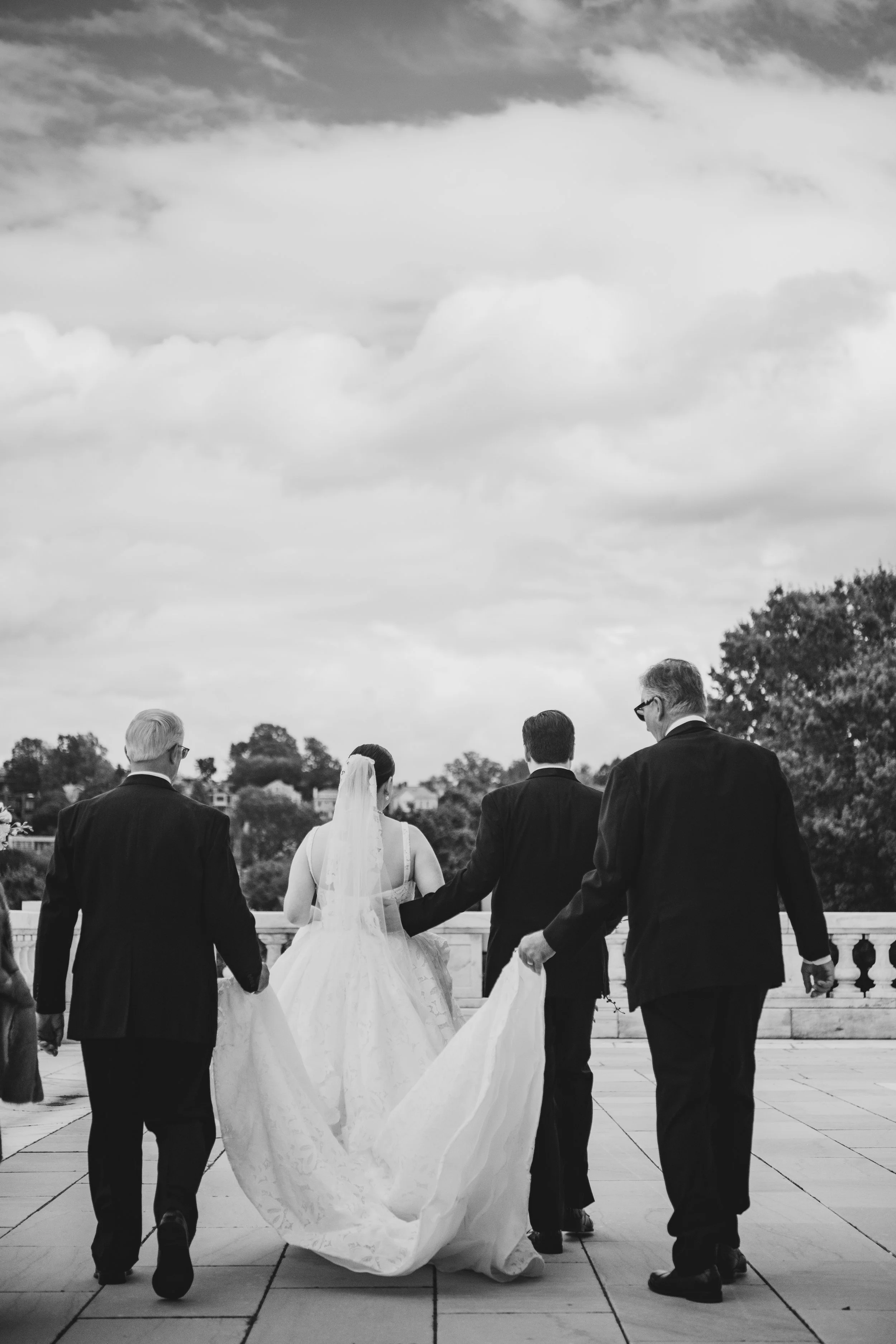 Black and white photo of a bride in a white wedding dress and veil walking with four men in dark suits, holding her gown as they walk on a terrace. The background features trees and a cloudy sky.