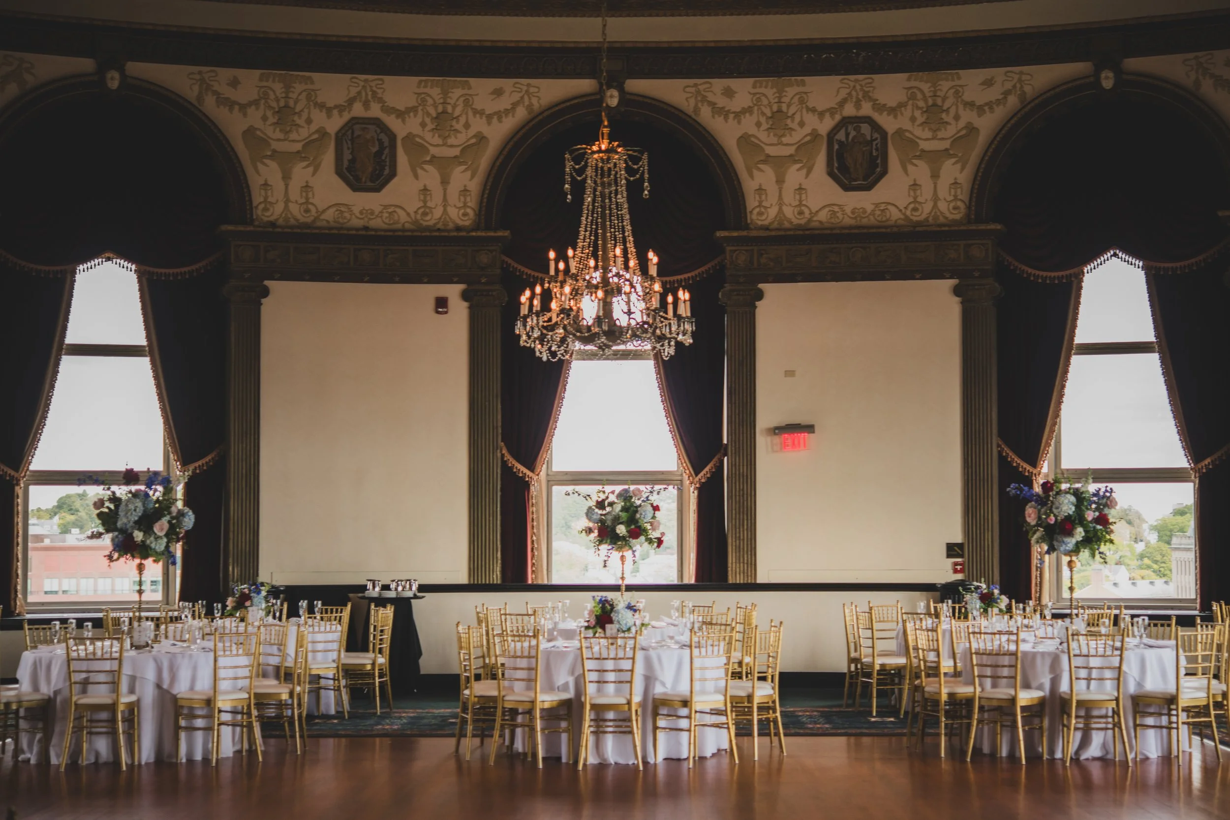 Elegant banquet hall with large windows, draped curtains, ornate ceiling, chandelier, and round tables with white tablecloths and gold chairs, decorated with floral centerpieces.