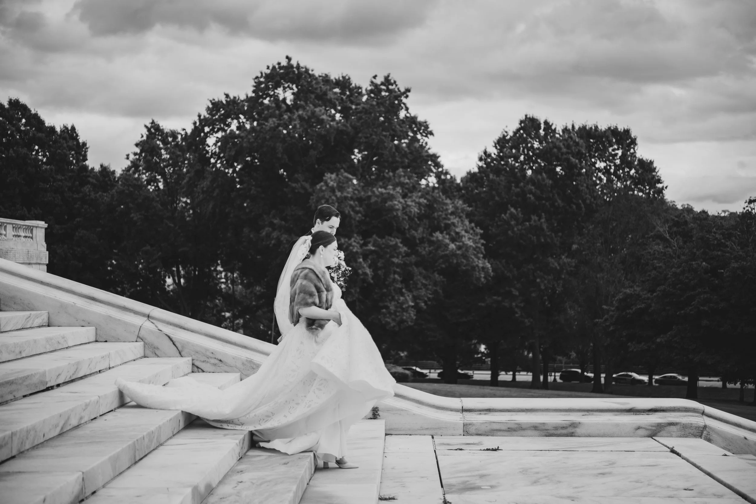 Black and white photo of a bride and groom walking down marble steps outdoors, with trees and cloudy sky in the background.